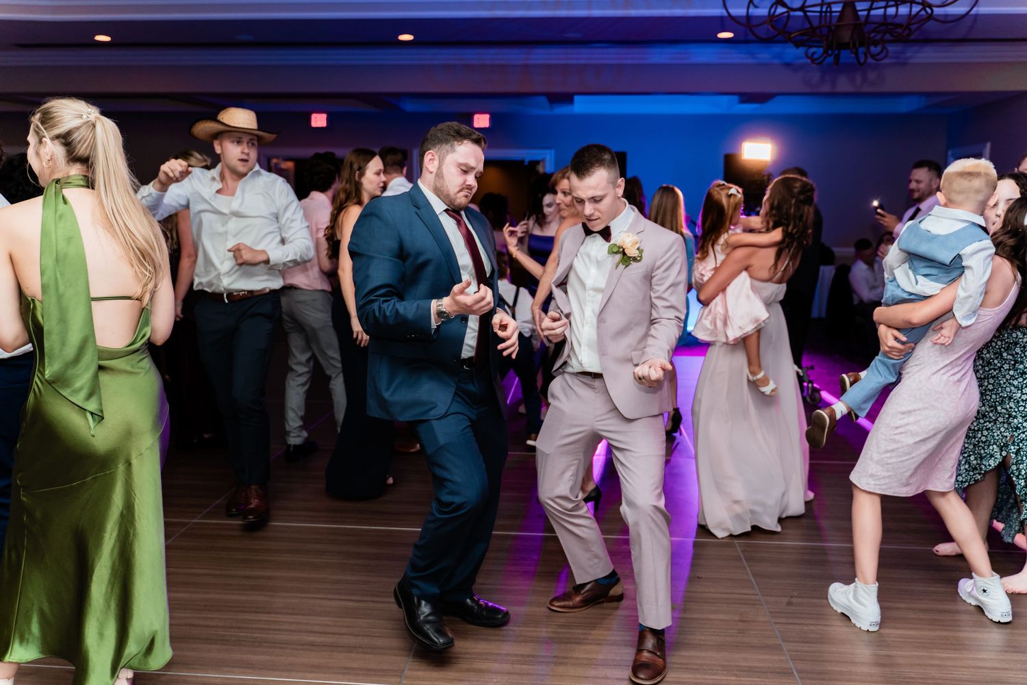 Wedding party guests dance and celebrate on a purple-lit dance floor during an energetic evening reception.