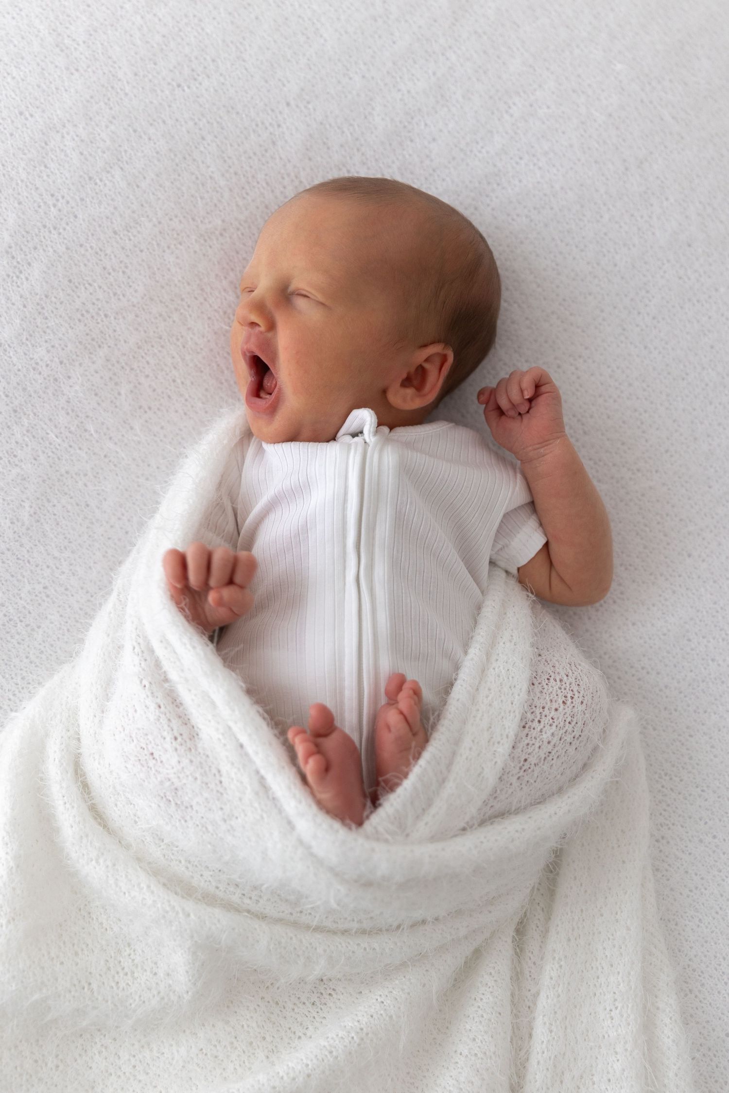 A yawning newborn baby wrapped in white blankets against a light background.