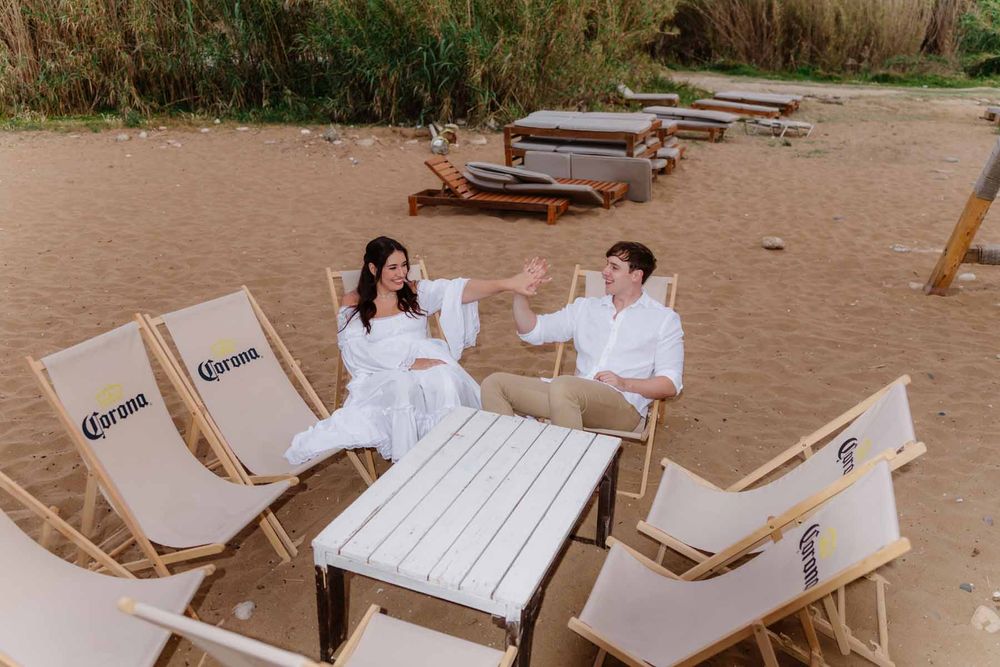 Engaged couple giving a high-five while sitting on beach loungers around a white table at Sandy Beach in Paphos, Cyprus, surrounded by sand