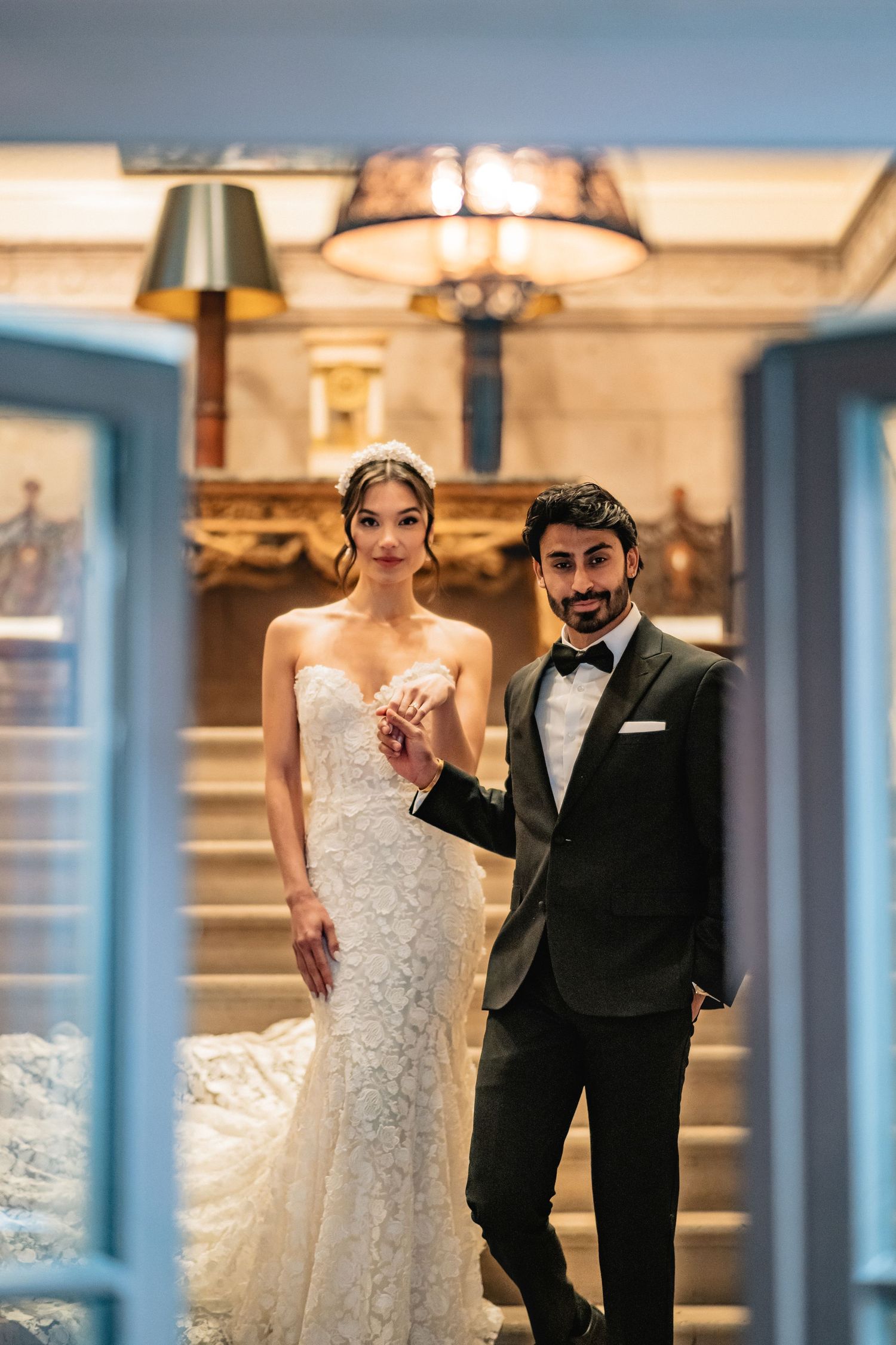 Elegant wedding couple stands in front of ornate interior with warm lighting and decorative fixtures.