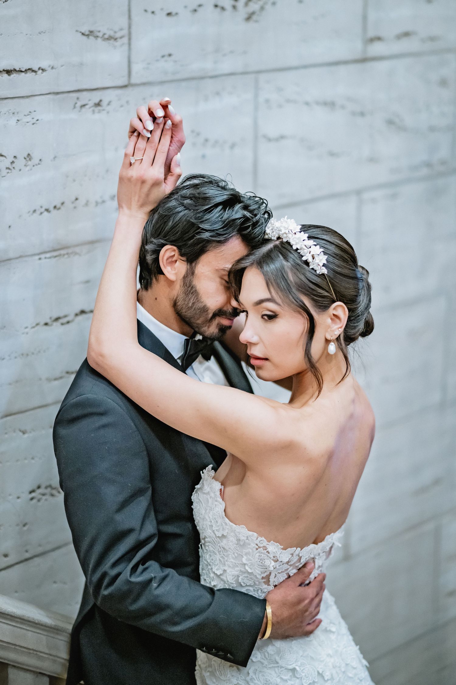 A romantic couple embraces in wedding attire against a concrete wall, with the bride wearing a delicate floral headpiece.