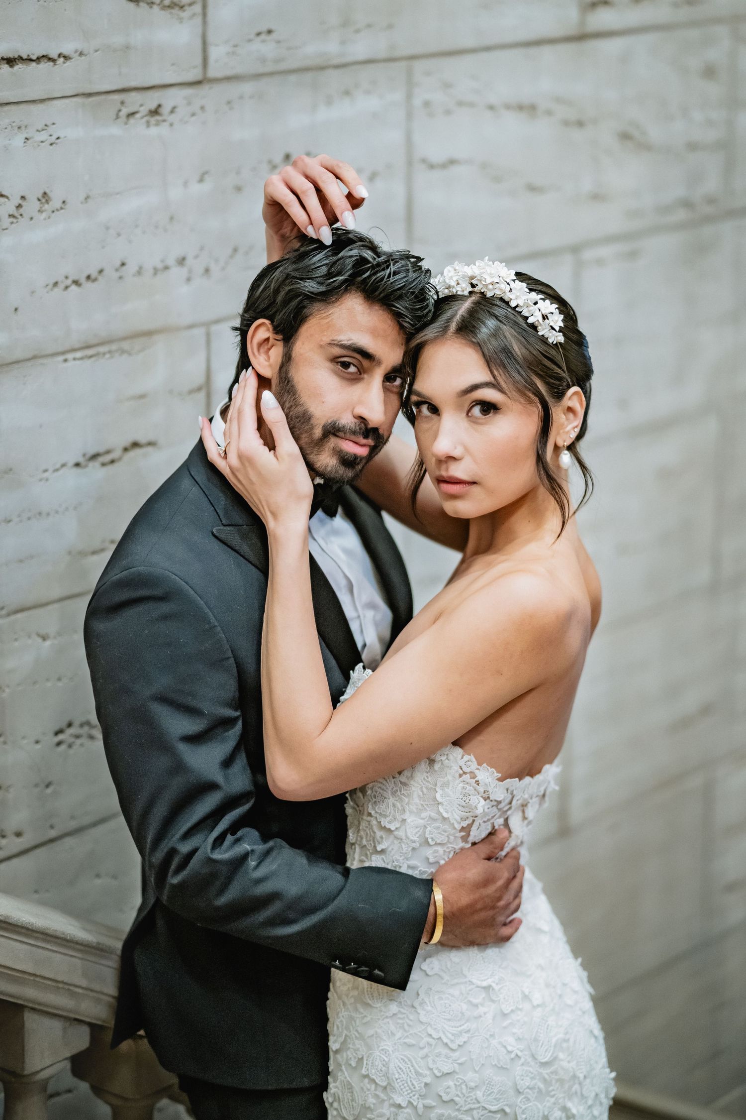 A romantic wedding portrait of a couple in formal attire embracing against a neutral wall in soft lighting.