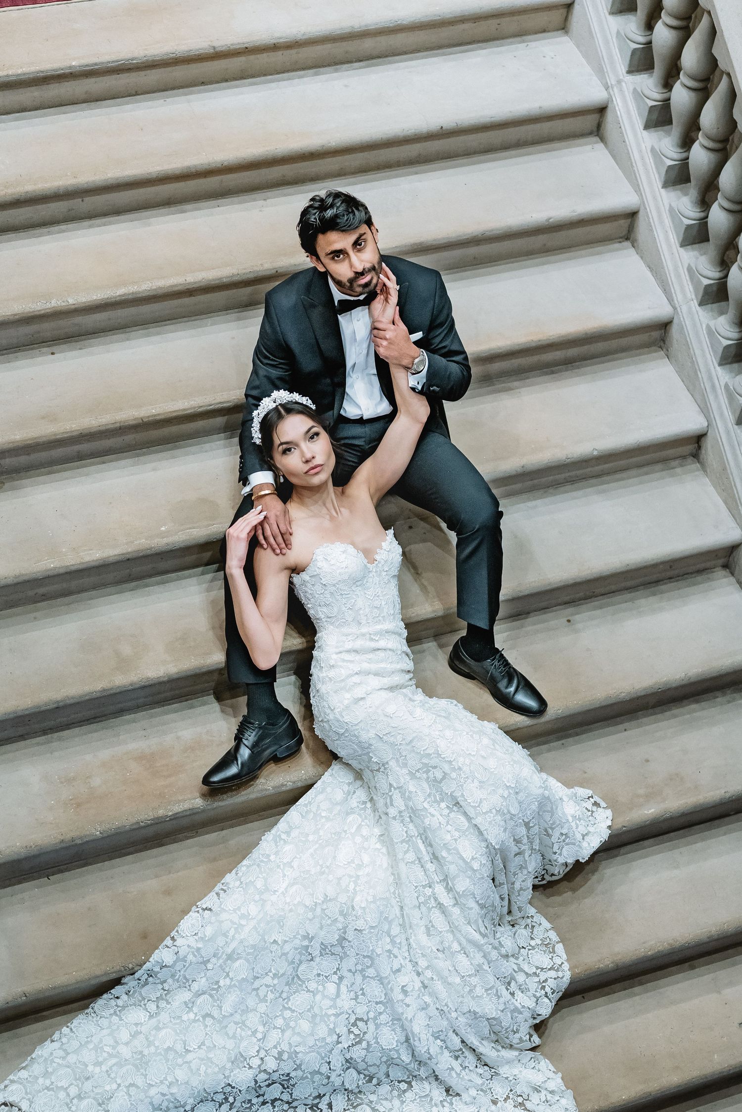 A couple in wedding attire poses playfully on a grand stone staircase, with the bride in a flowing lace gown.
