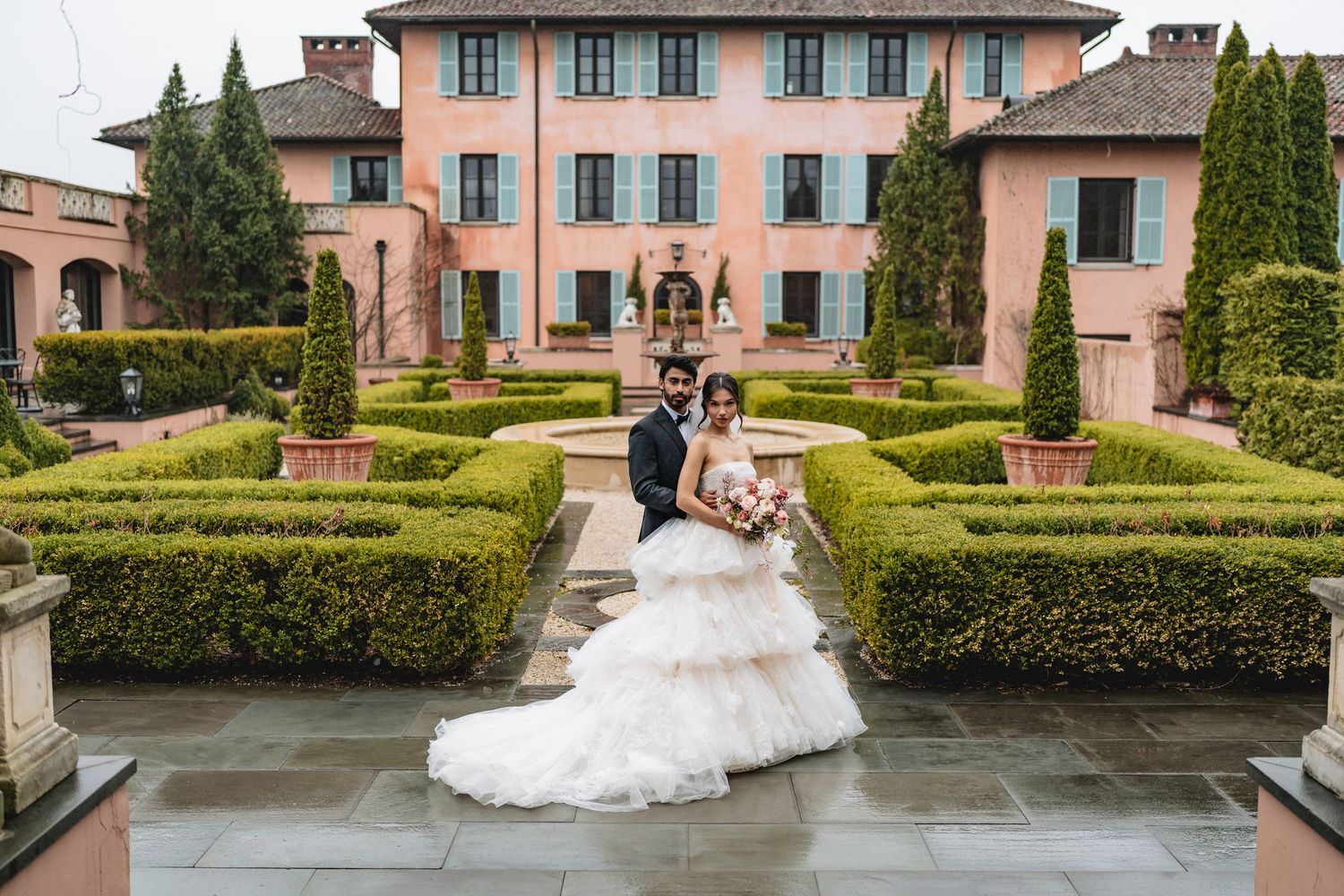 A couple poses in formal wedding attire in front of an elegant pink Italian villa with manicured topiary gardens.