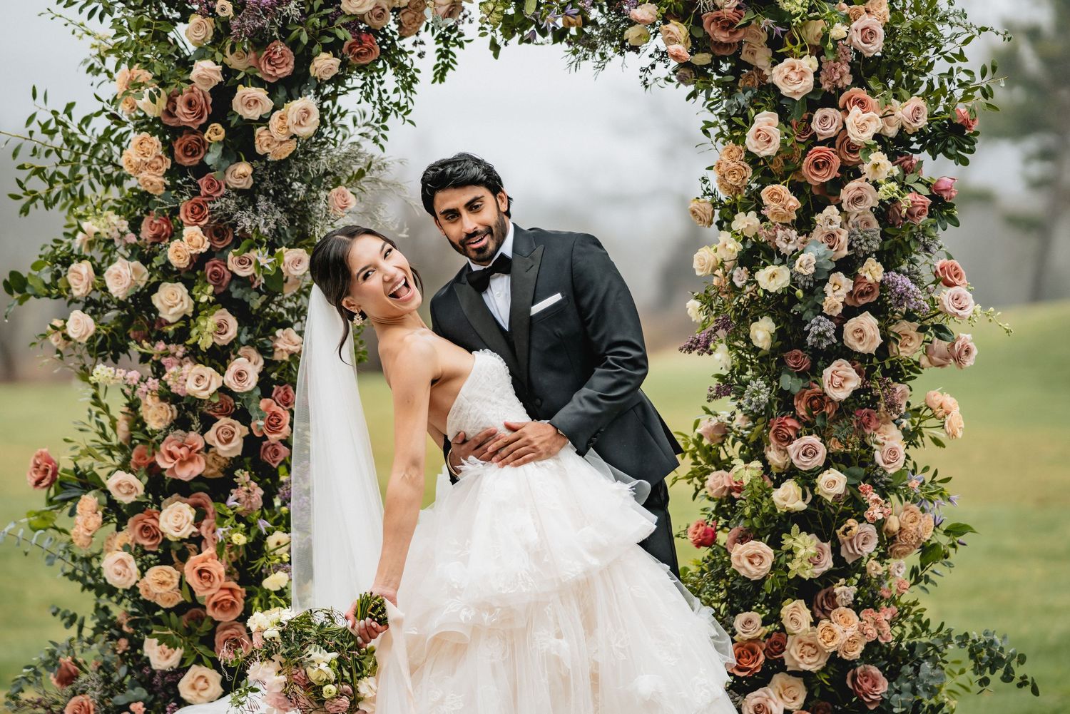 A wedding couple poses beneath an ornate floral arch with peach and pink roses in an outdoor garden setting.