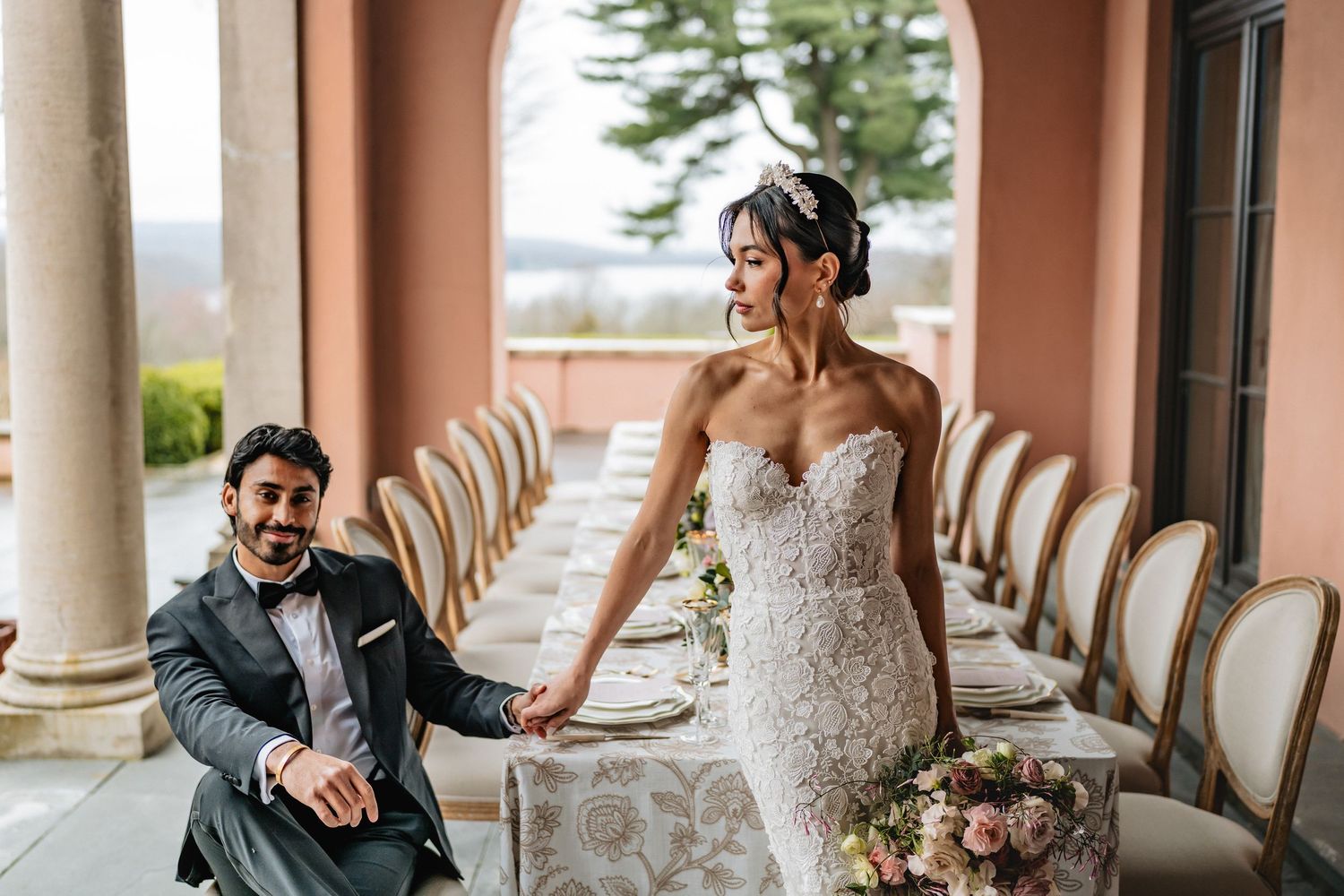 A bride in a lace strapless wedding gown and groom in a tuxedo pose together in an elegant columned venue.