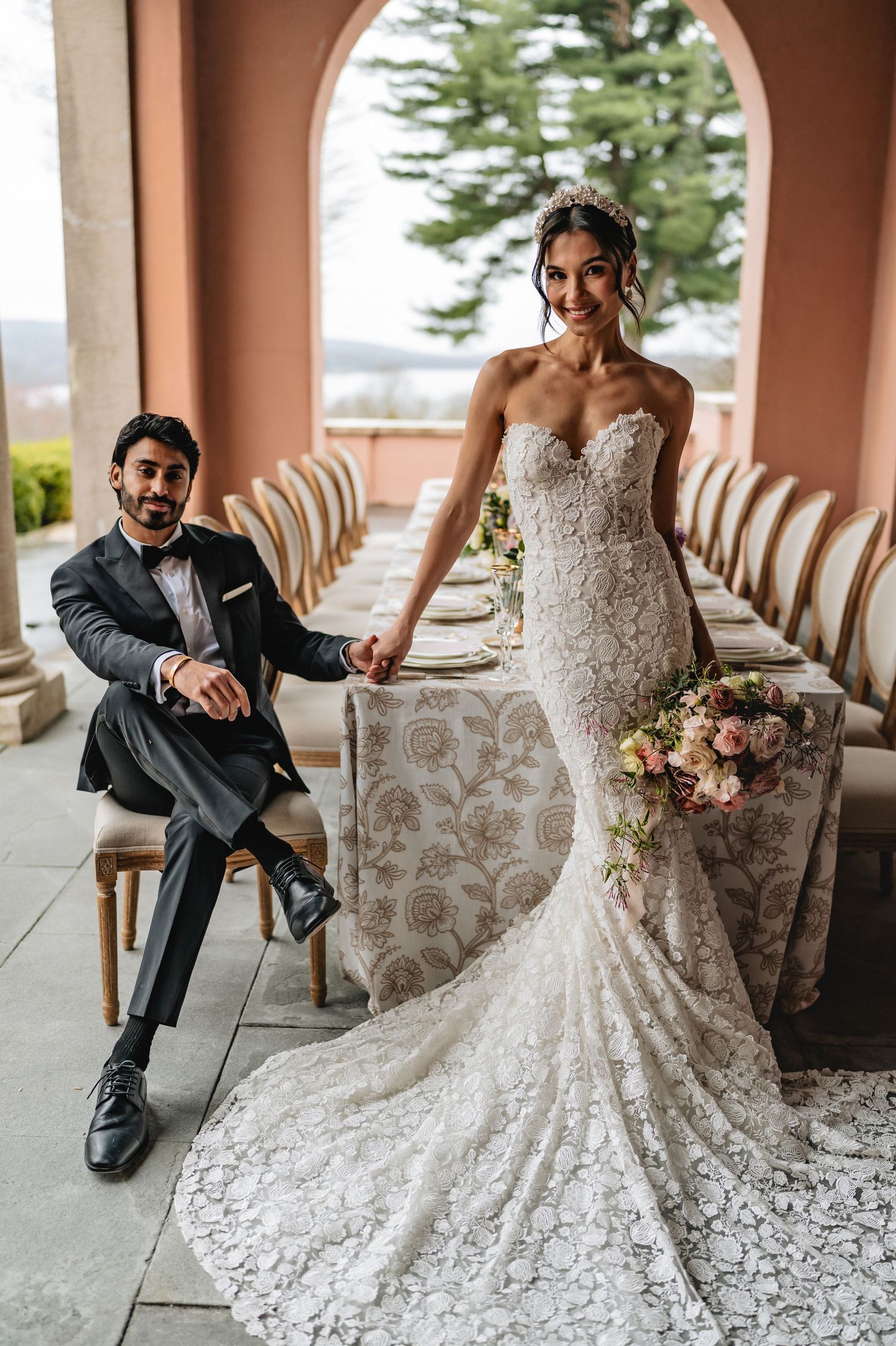 Elegant bride in lace mermaid wedding dress poses with groom seated in antique chair on covered terrace.