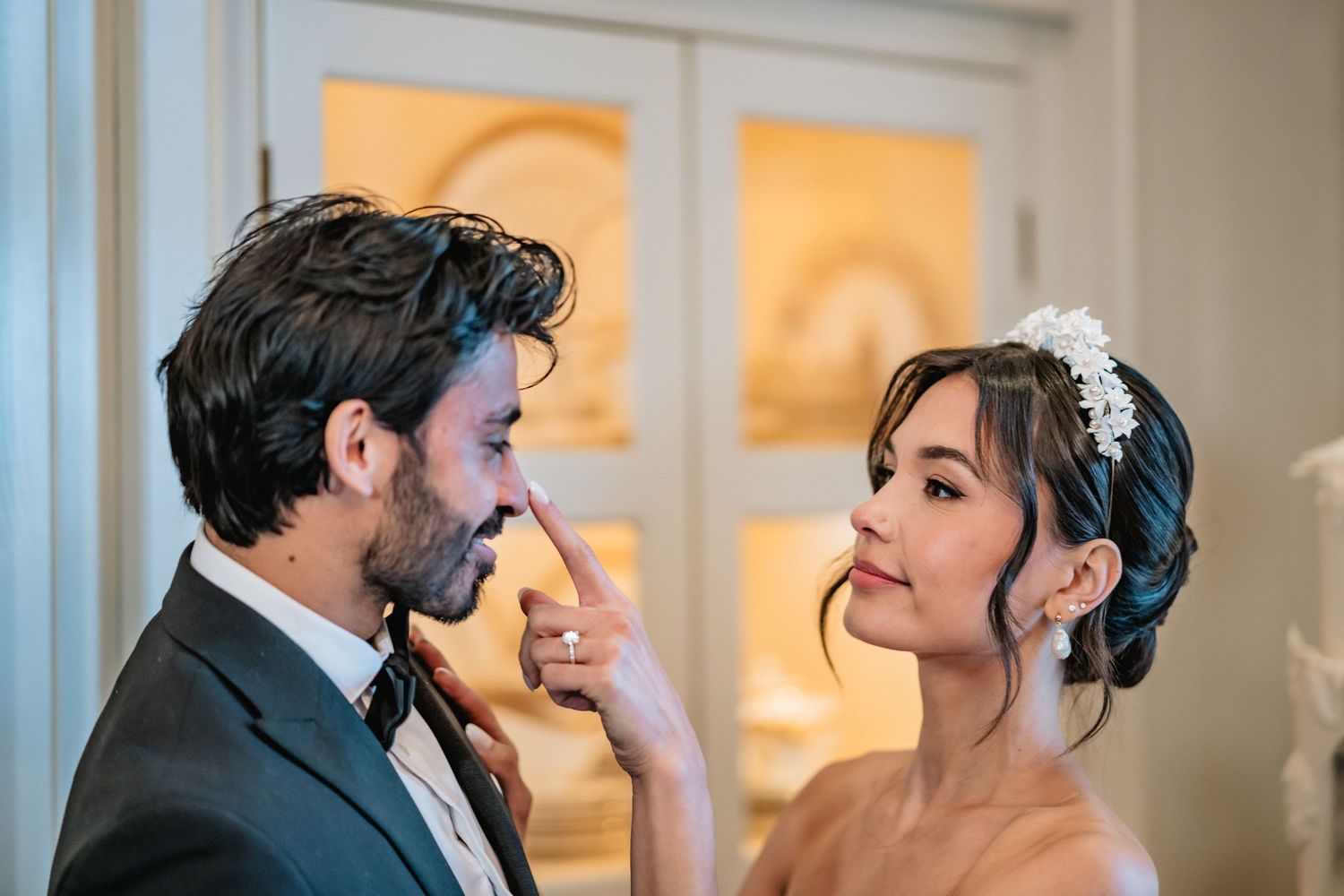 A couple shares an intimate romantic moment on their wedding day, with warm lighting glowing through windows in the background.