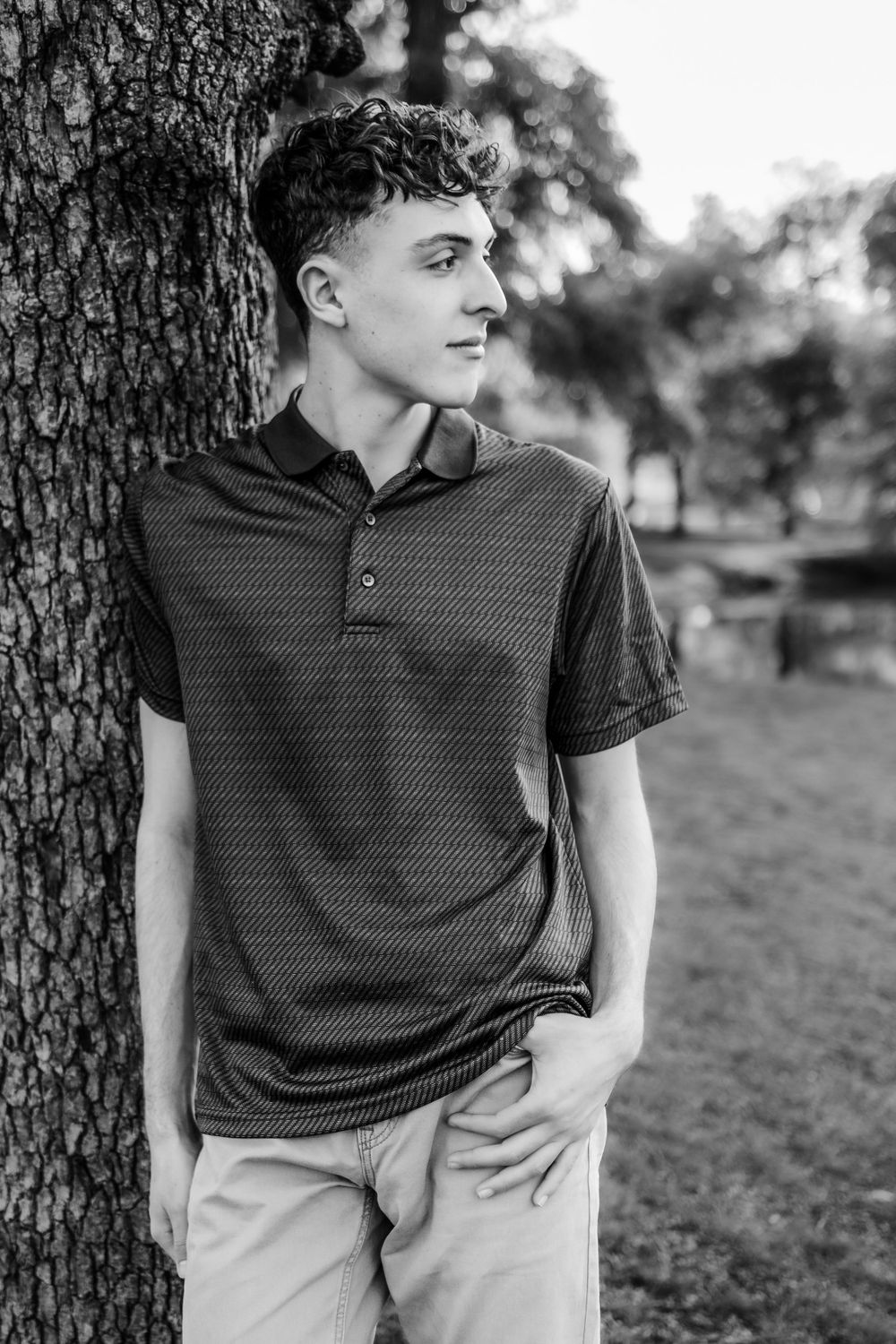 Black and white portrait of person in polo shirt leaning against tree trunk in park setting.