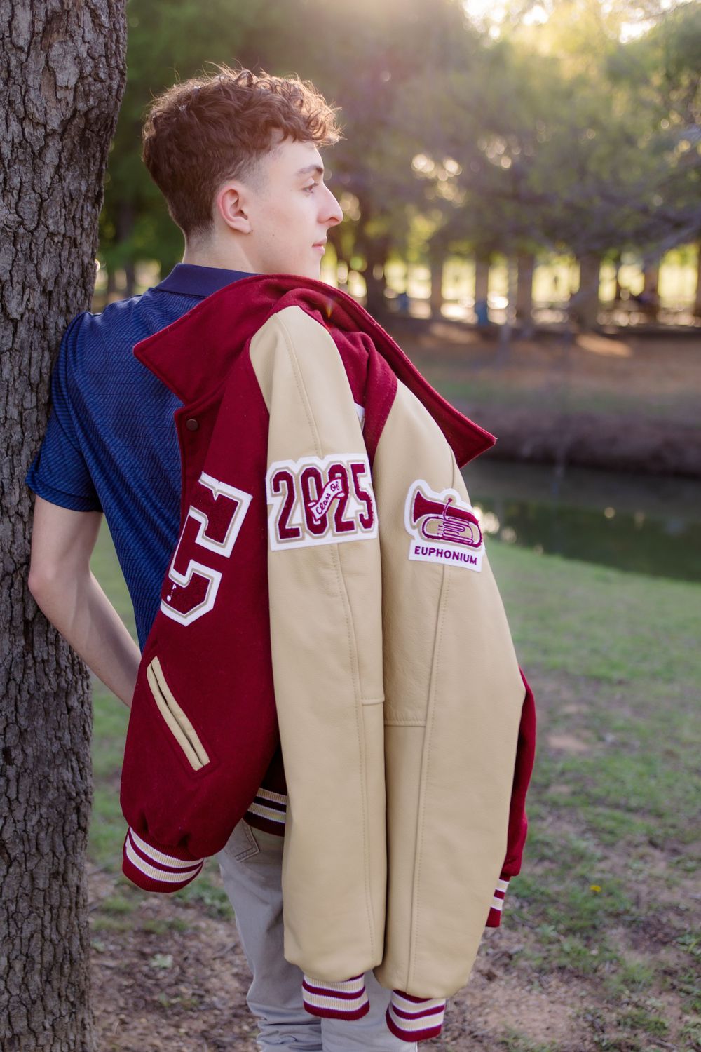 Student wearing varsity letterman jacket with class year 2025 poses by tree in outdoor setting at sunset.