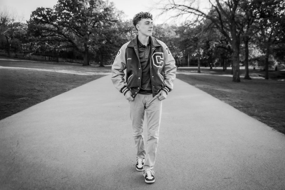 A black and white photo of someone wearing a varsity jacket and jeans walking on a tree-lined path.