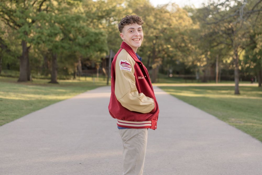 A person wearing a red and beige varsity jacket poses on a tree-lined path during golden hour.
