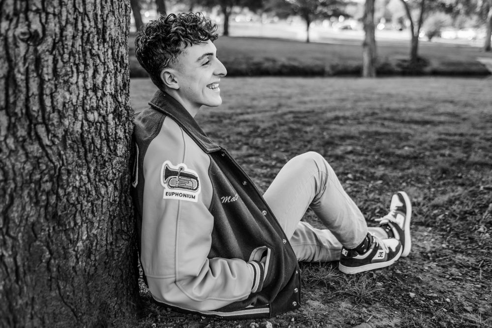 A young athlete in a varsity jacket sits relaxing against a tree trunk in black and white photography.