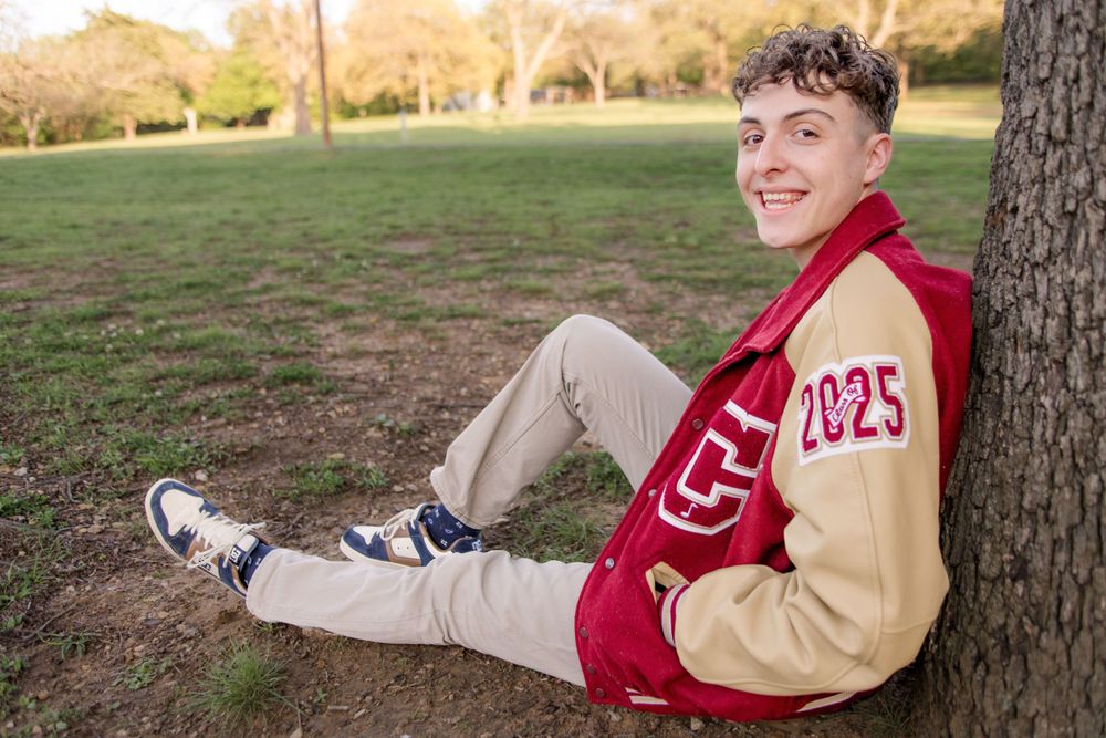 Young student in red and tan varsity jacket sits casually against tree in sunny park setting.