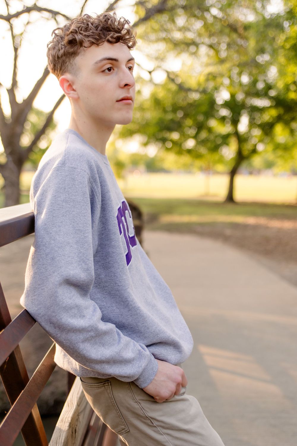 A person in a gray sweatshirt leans against a park bench on a sunny autumn day with trees in the background.