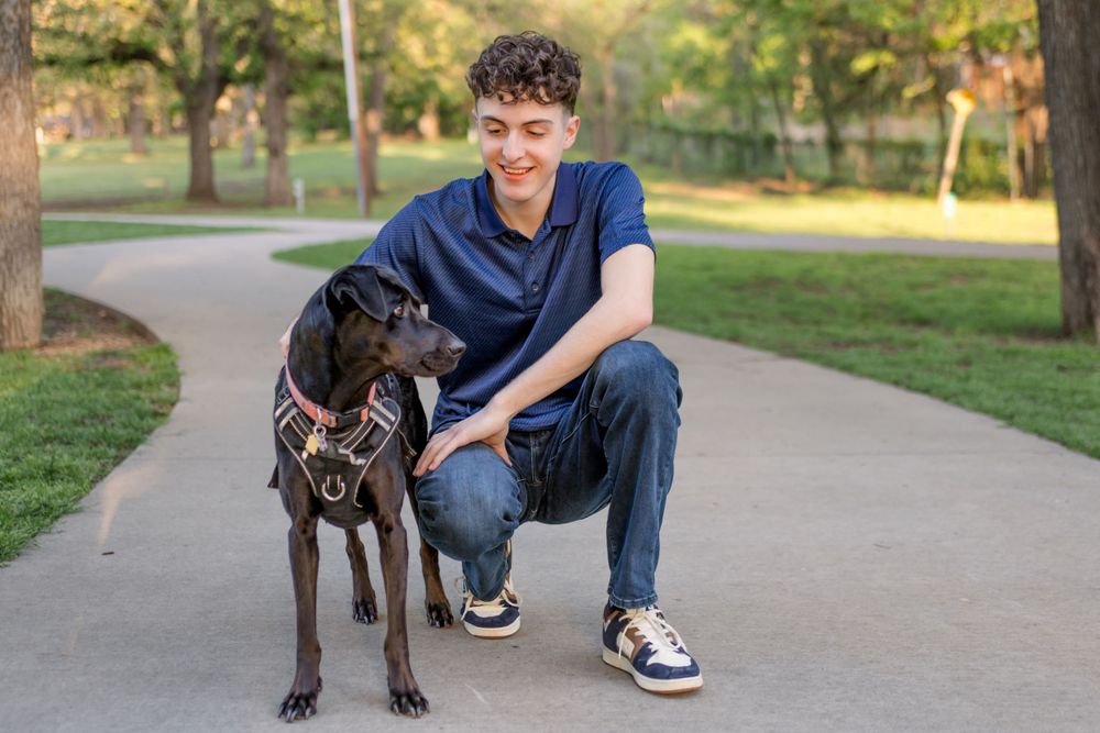 Someone in a navy blue shirt crouches next to their friendly black dog on a walking path in a sunny park.