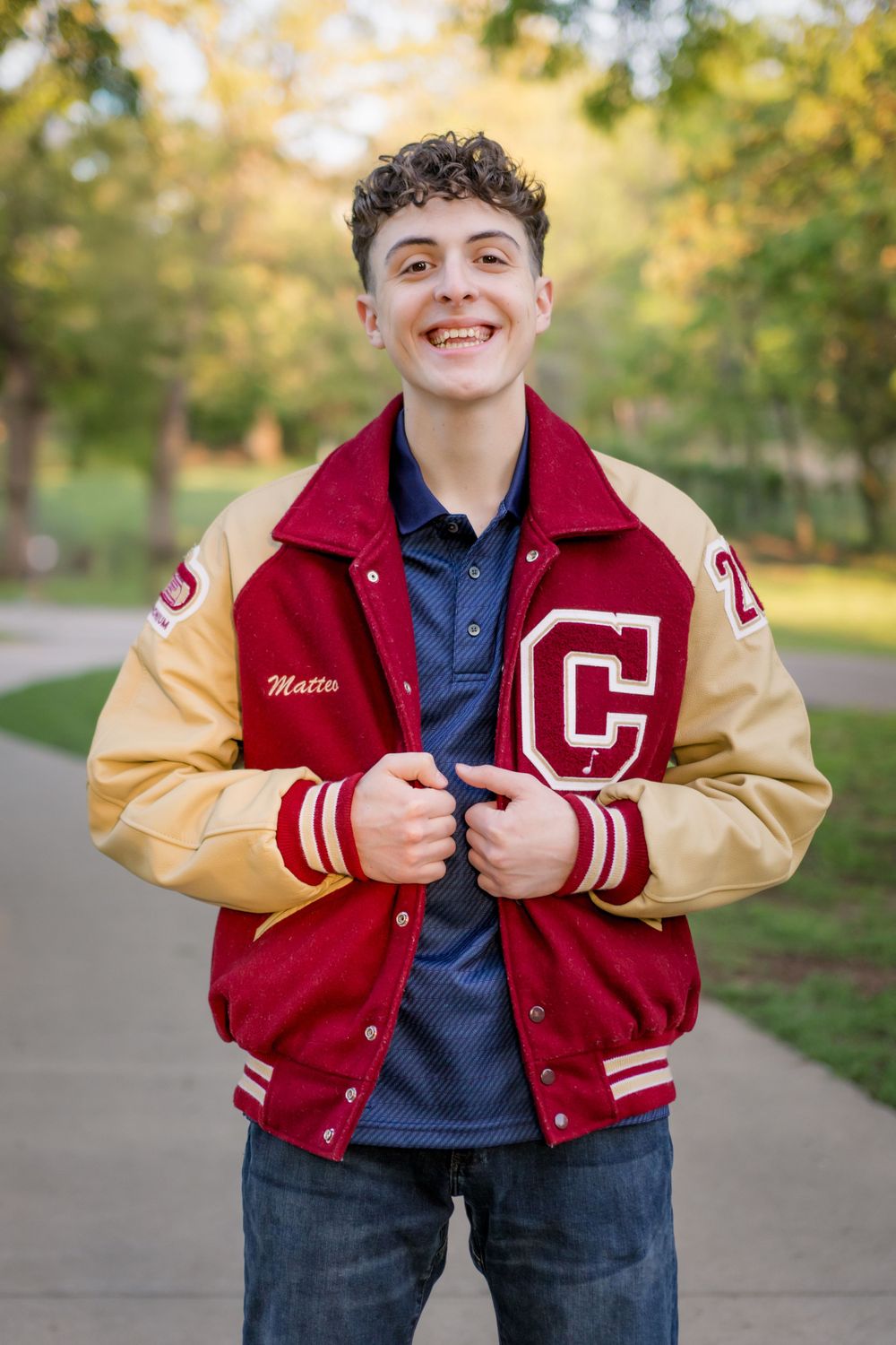 A student wearing a red and gold varsity letterman jacket with a C letter poses outdoors in a park.