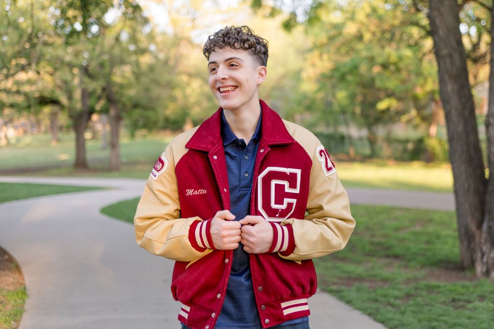 Student smiling outdoors wearing red and tan varsity letterman jacket with C logo on park pathway in autumn setting.