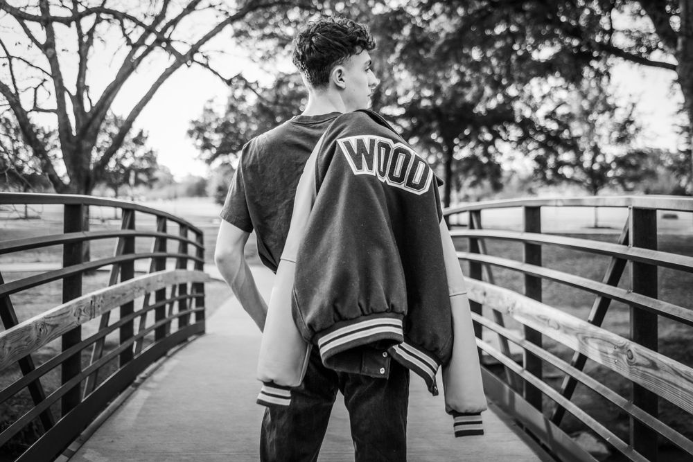 Black and white photo of someone in a varsity jacket standing on a wooden bridge.