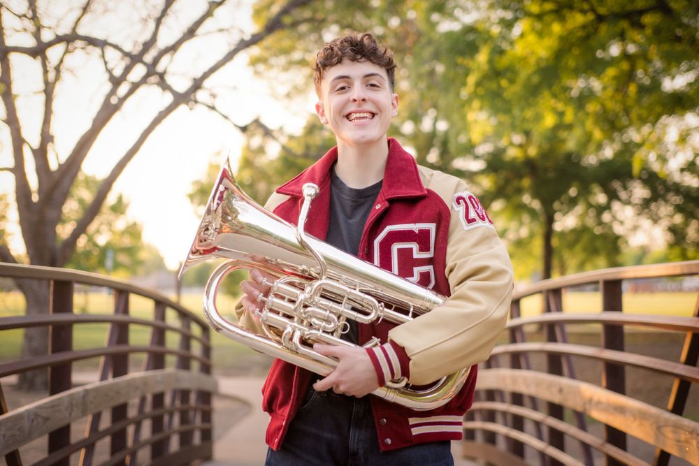 Student musician poses with brass euphonium instrument on wooden bridge during golden hour.