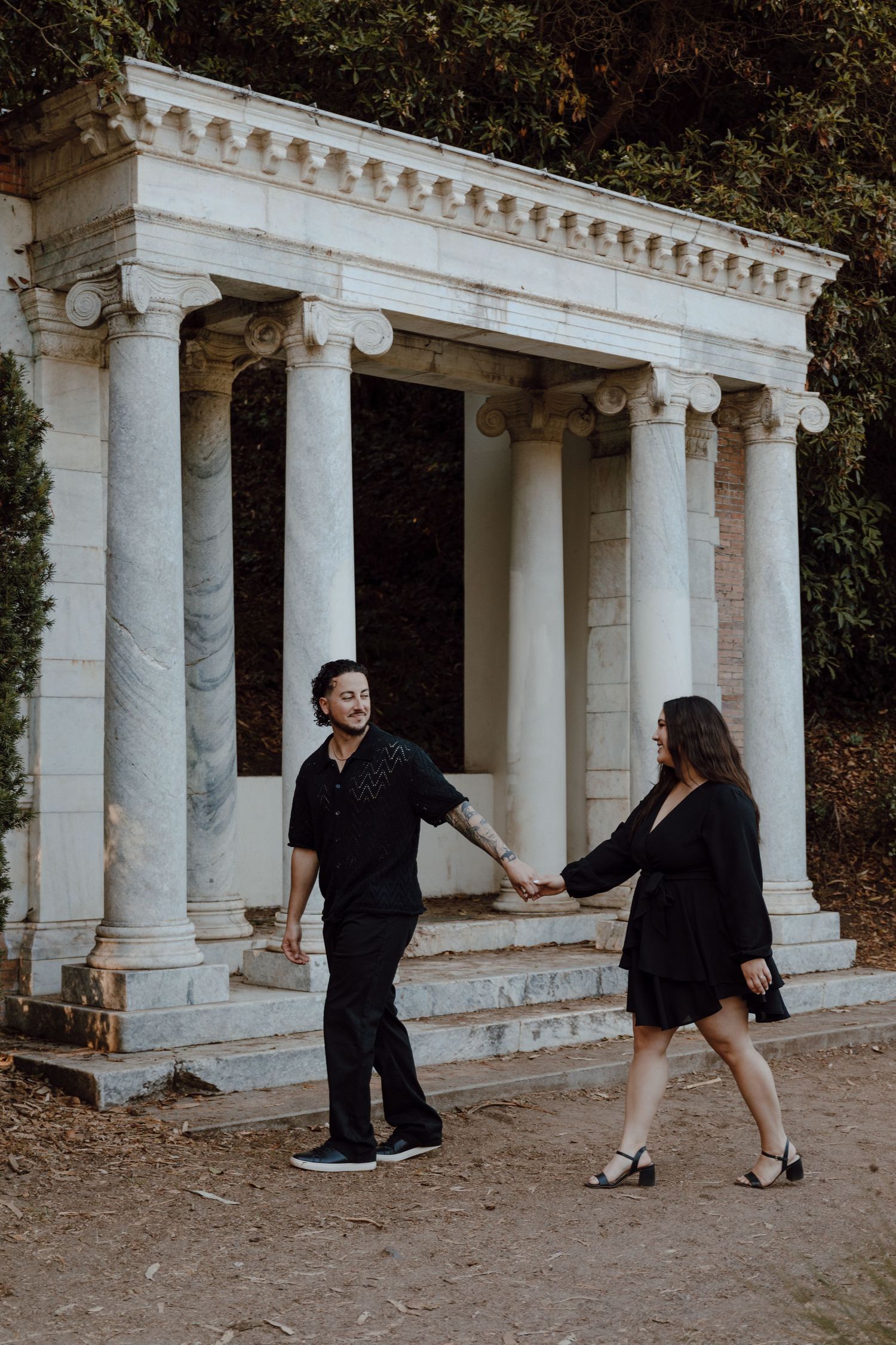 A couple holds hands and dances near an elegant white marble colonnade in a garden setting at sunset.