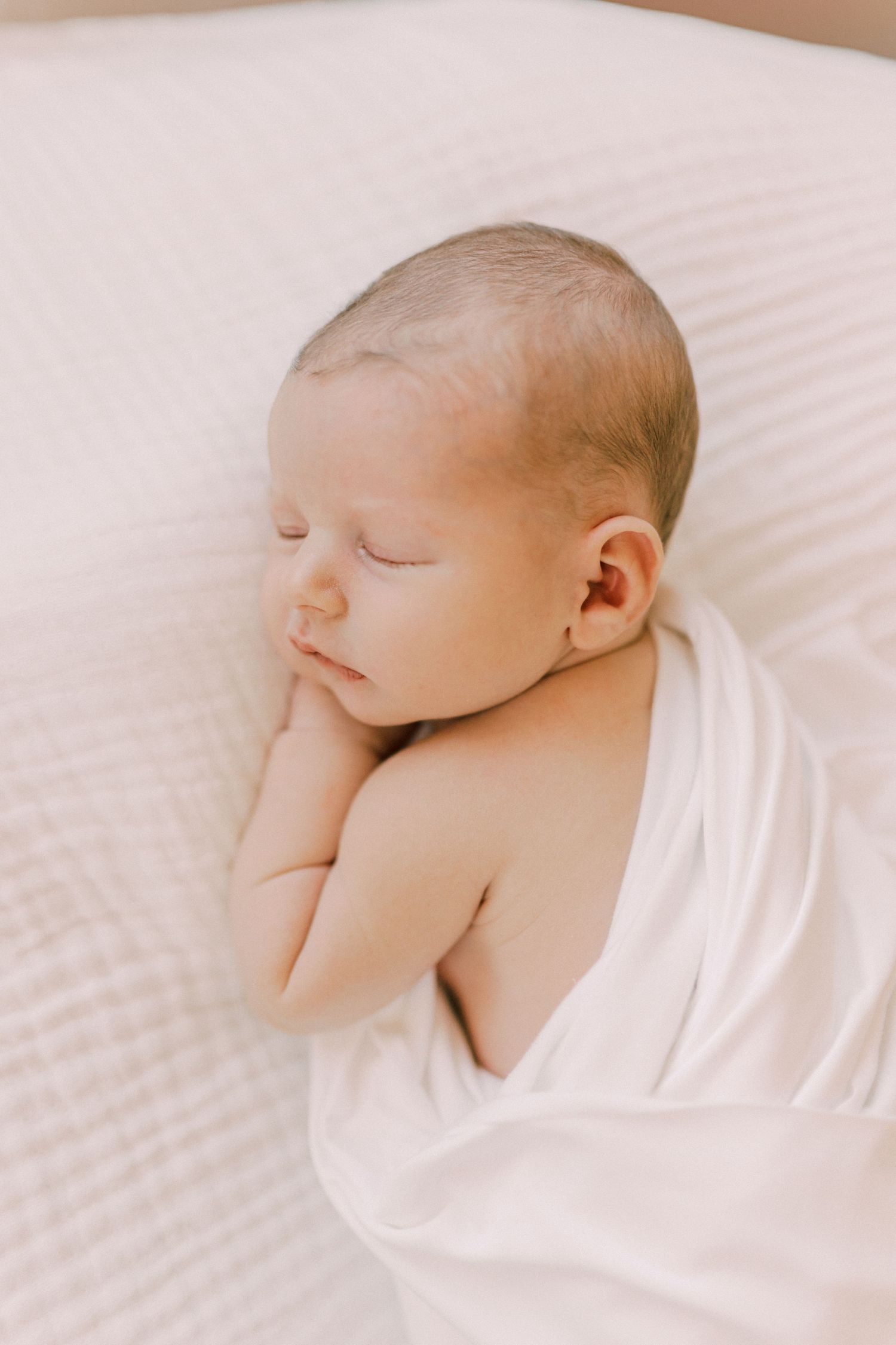 Peaceful sleeping newborn wrapped in soft white blanket resting on textured white bedding.