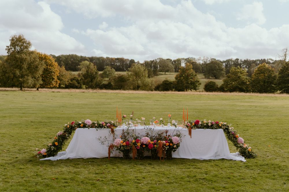 An outdoor wedding setup in a green field with a white altar decorated with orange and pink floral arrangements.