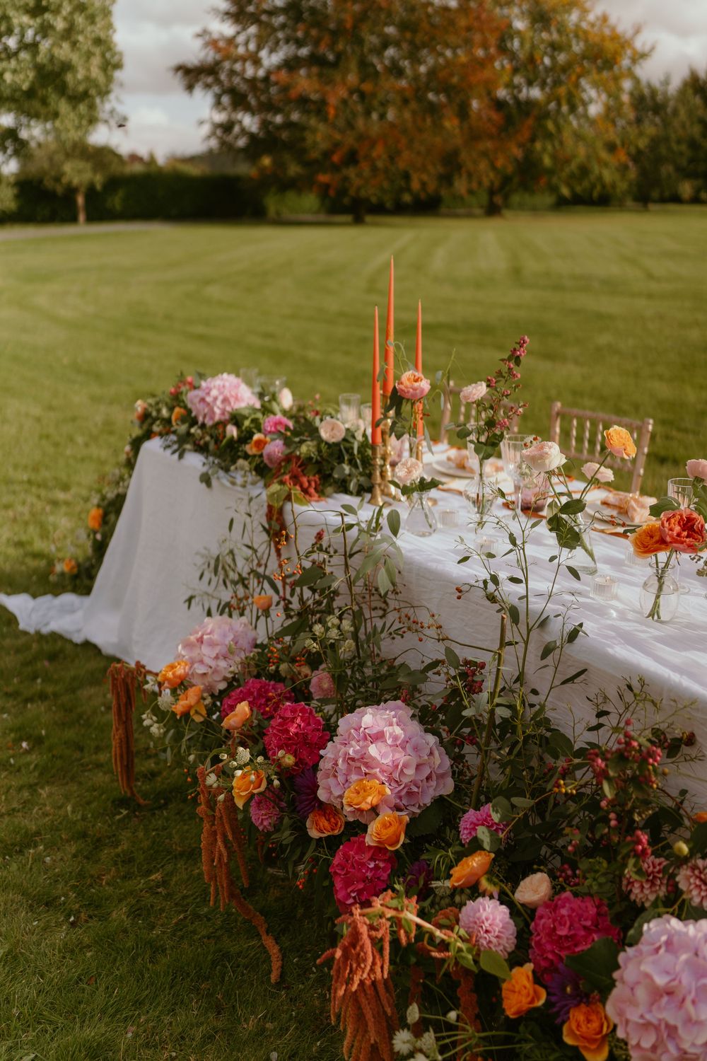 Elegant outdoor table setting with lush floral arrangements featuring pink and red blooms against an autumn garden backdrop.