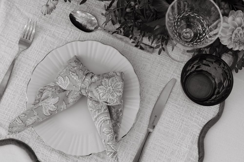 Overhead view of a table setting with a paper plate, cutlery, and dark glass against a white tablecloth.
