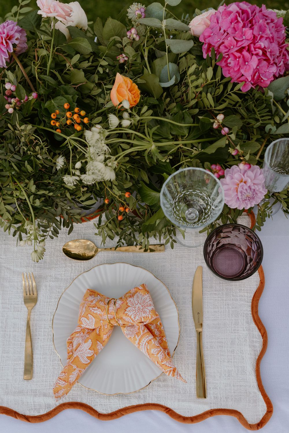 Elegant table setting with pink floral arrangements, white linen tablecloth, and decorative orange napkins folded in bow shape.