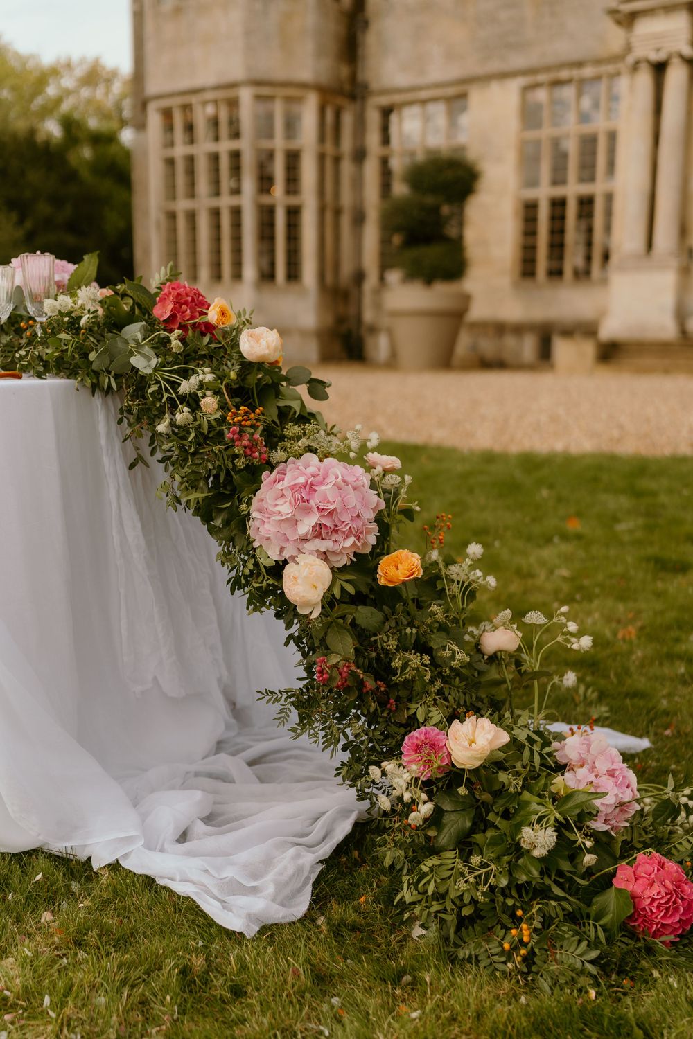 White-clothed table adorned with cascading pink and red flowers beside a historic stone mansion with large windows.