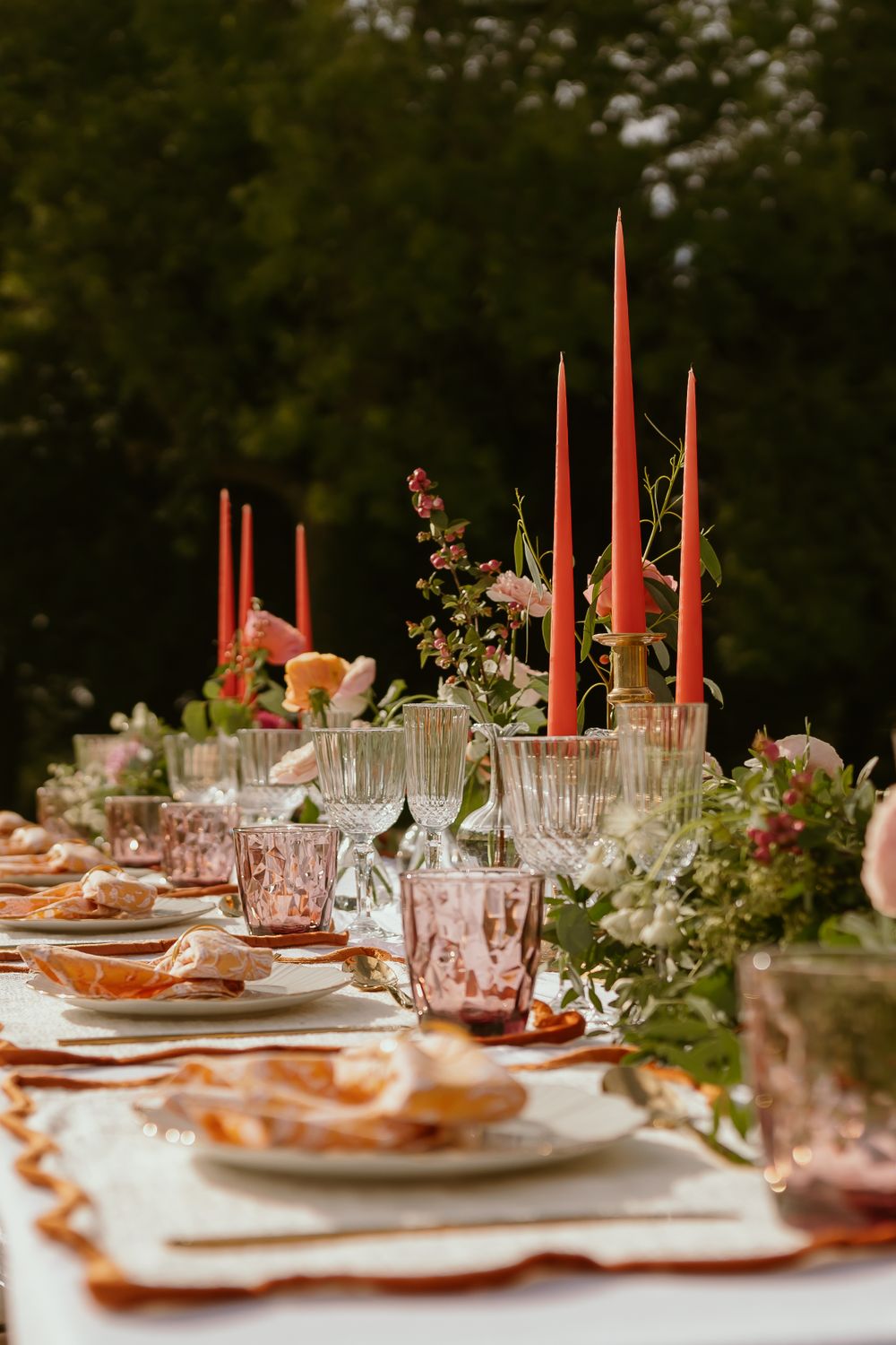Elegant table setting with coral taper candles, pink glassware, and greenery garland against dark moody background.