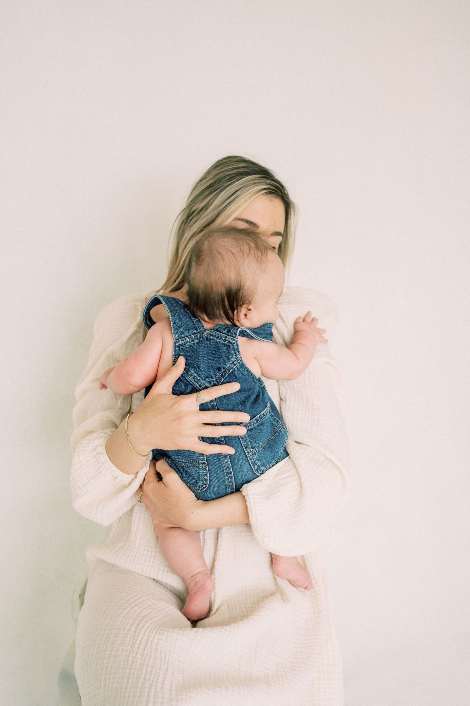A tender moment captured as a mother in a cream sweater holds an infant wearing blue denim overalls.