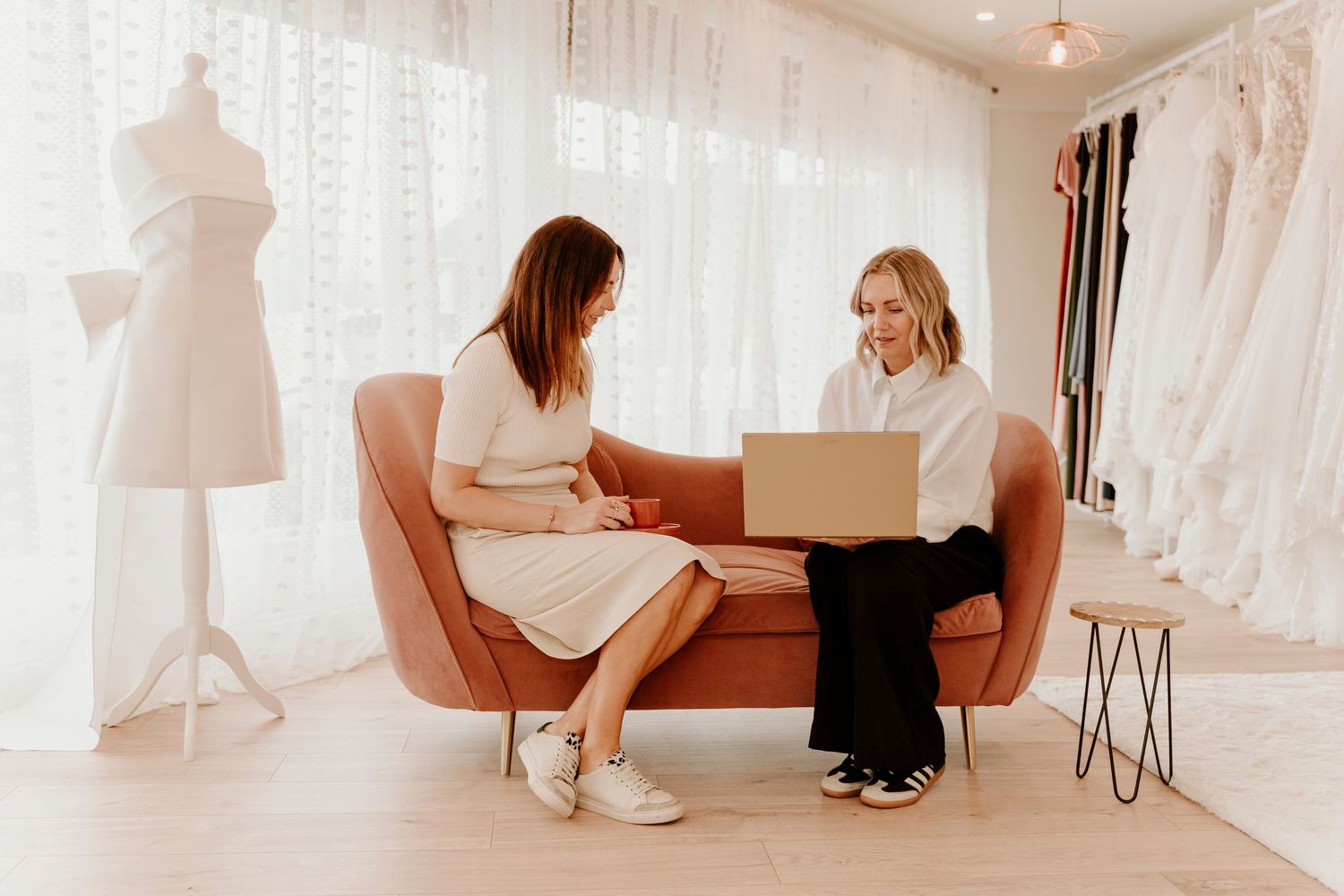 Two people sit on a peach-colored couch reviewing a laptop in a bridal boutique with white dresses in the background.