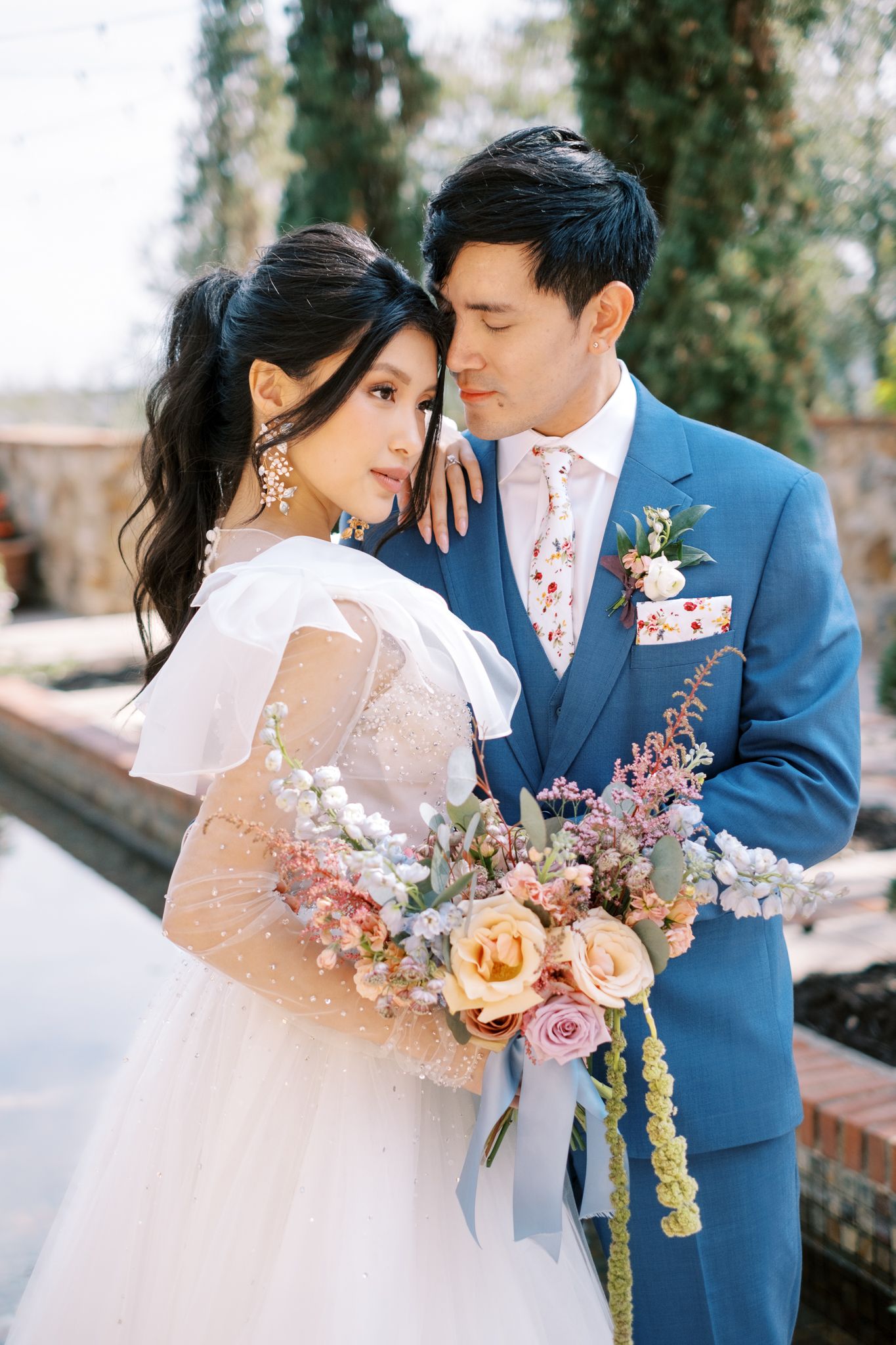 Romantic wedding portrait with couple holding stunning pastel floral bouquet against stone backdrop.