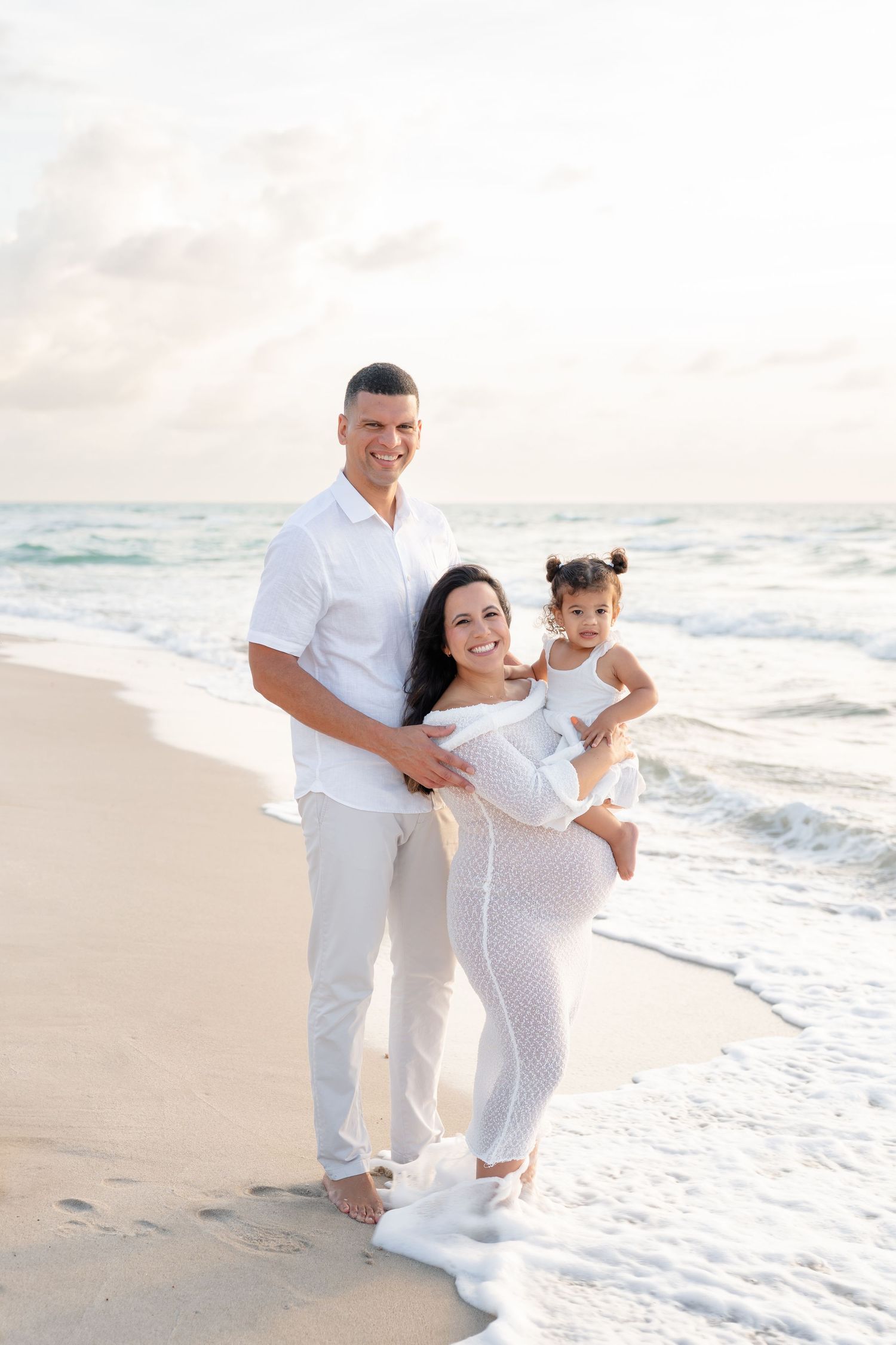 Expecting couple walking barefoot on beach near lighthouse during sunset maternity photo session.