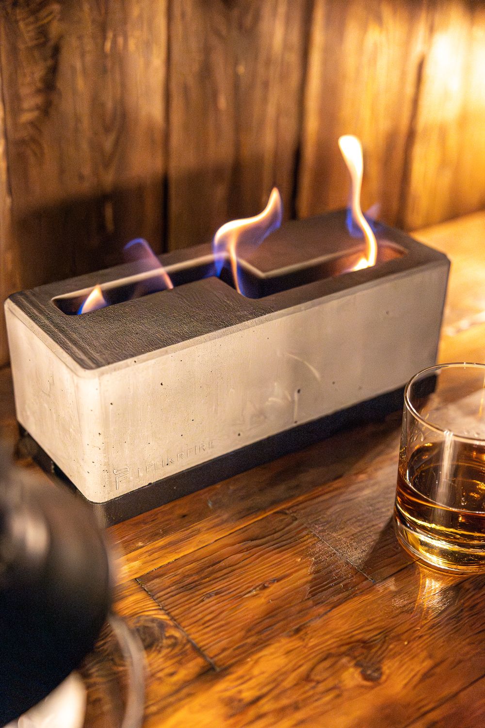 Concrete tabletop fire pit with blue flames next to glass of whiskey.