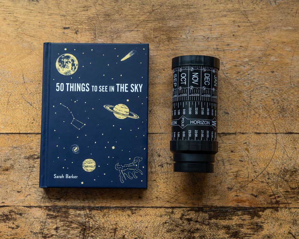 Book titled '50 Things to See in the Sky' next to black cylindrical container on wooden surface.