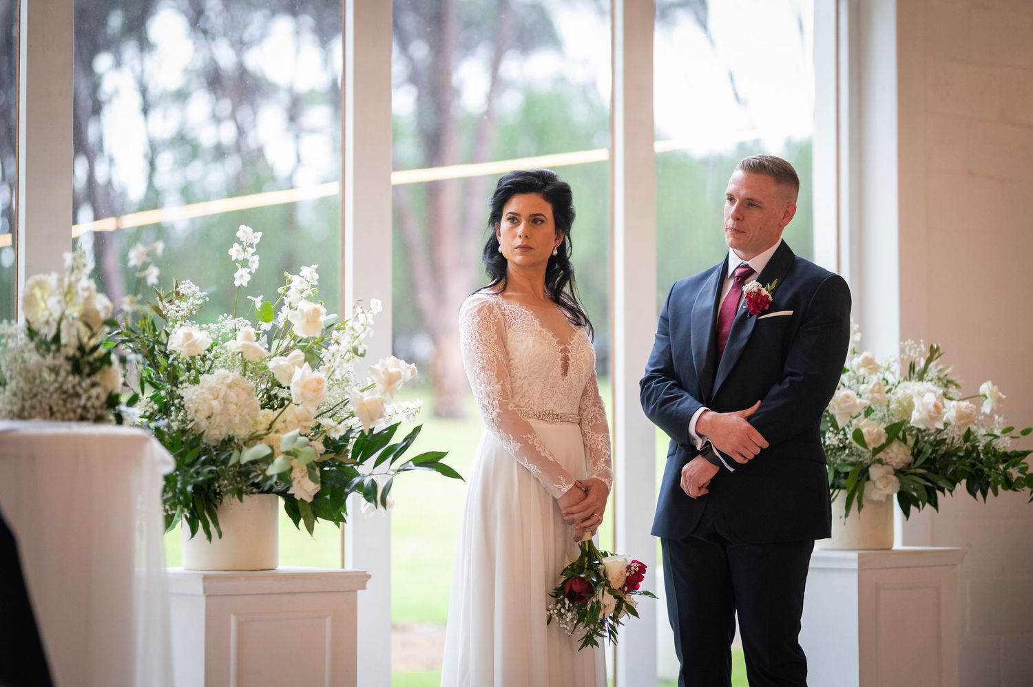 A wedding ceremony takes place in a bright chapel with an arched window and floral arrangements on pedestals at the altar.