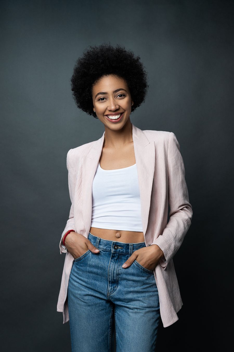 A person wearing a light pink blazer, white crop top and blue jeans poses confidently against a dark background.