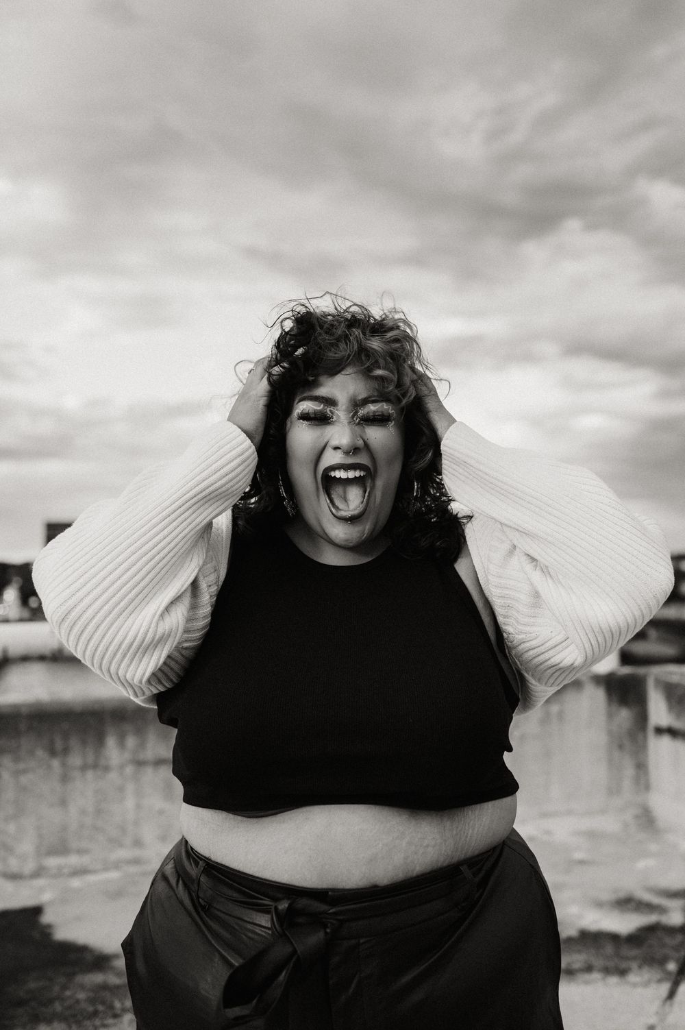 Black and white portrait of a joyful plus size woman laughing expressively in casual attire against a cloudy sky.