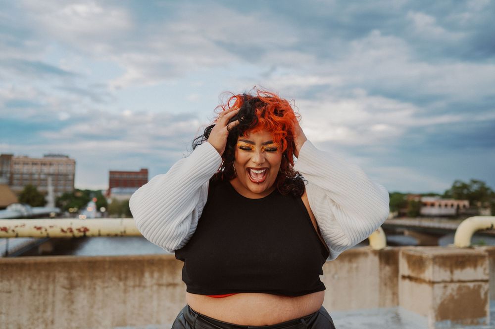 A joyful person with bright red hair wearing a black crop top poses expressively against a cloudy sky on a rooftop.