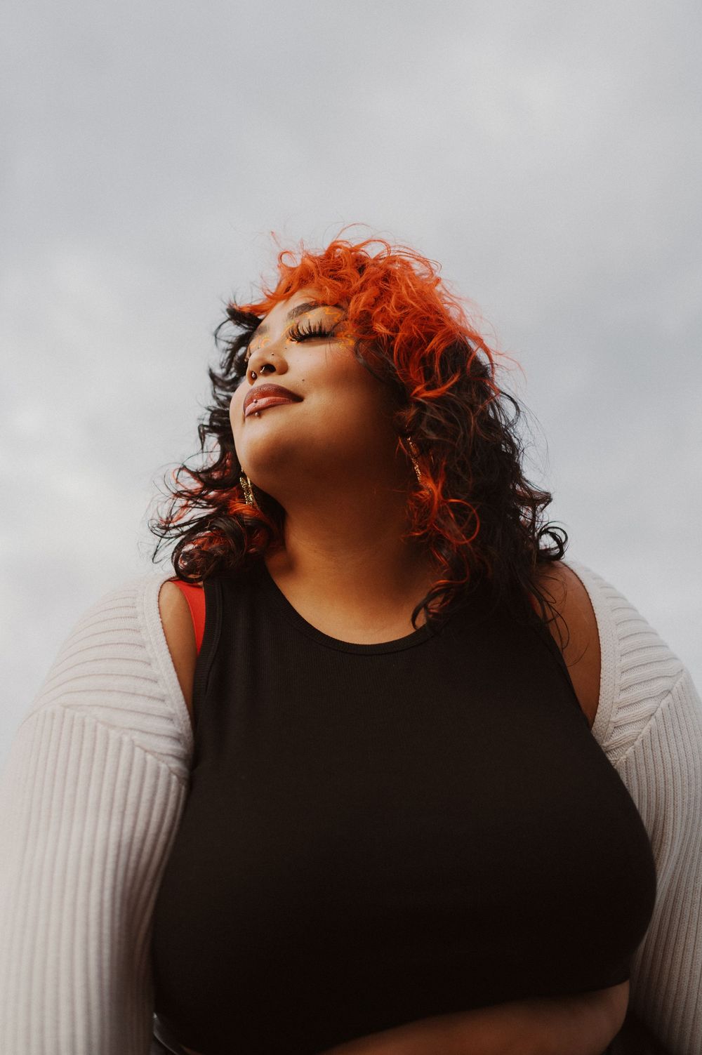 A person with bright red and dark curly hair tilts their head back against a moody gray sky while wearing a black top.