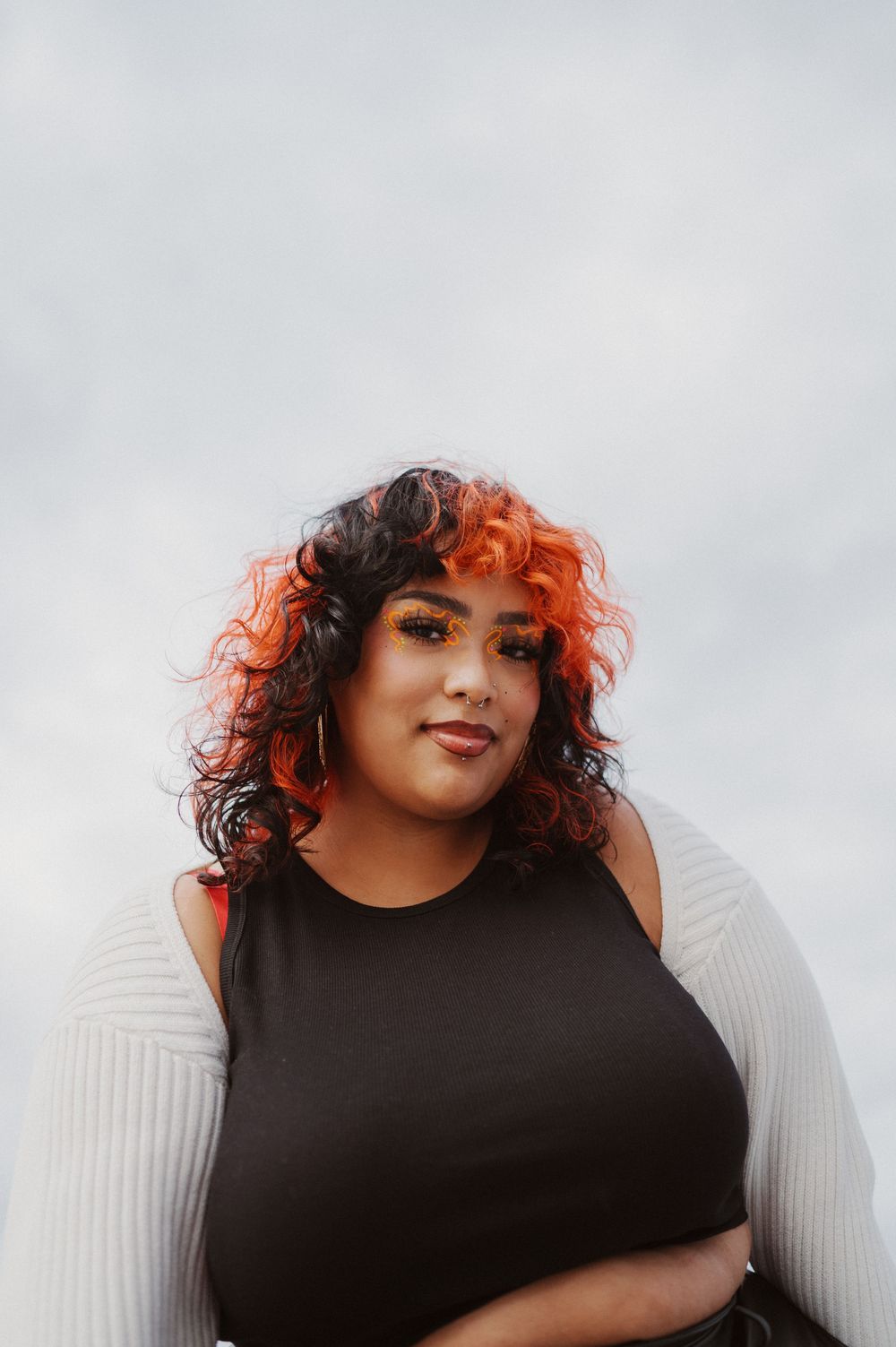 A portrait shot against a light background shows curly red and dark hair styled naturally with a black sleeveless top.