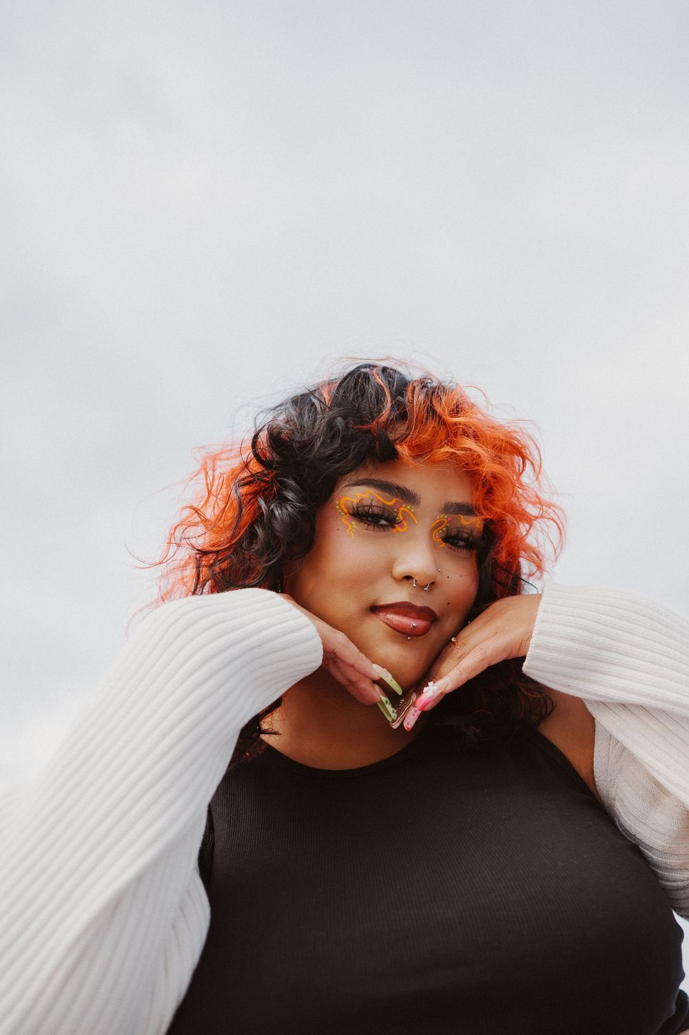 A fashion portrait with bright orange and black curly hair wearing a black top and white sweater against a light background.