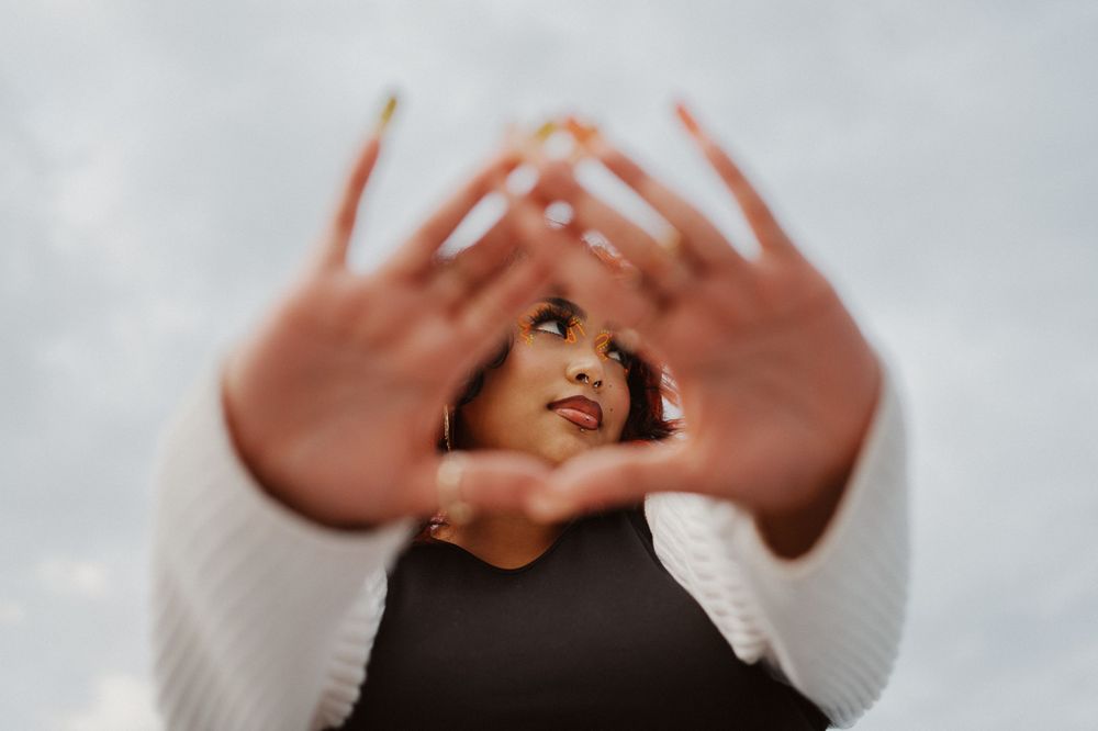 A person wearing white sleeves forms a heart shape with their hands against a cloudy sky background.