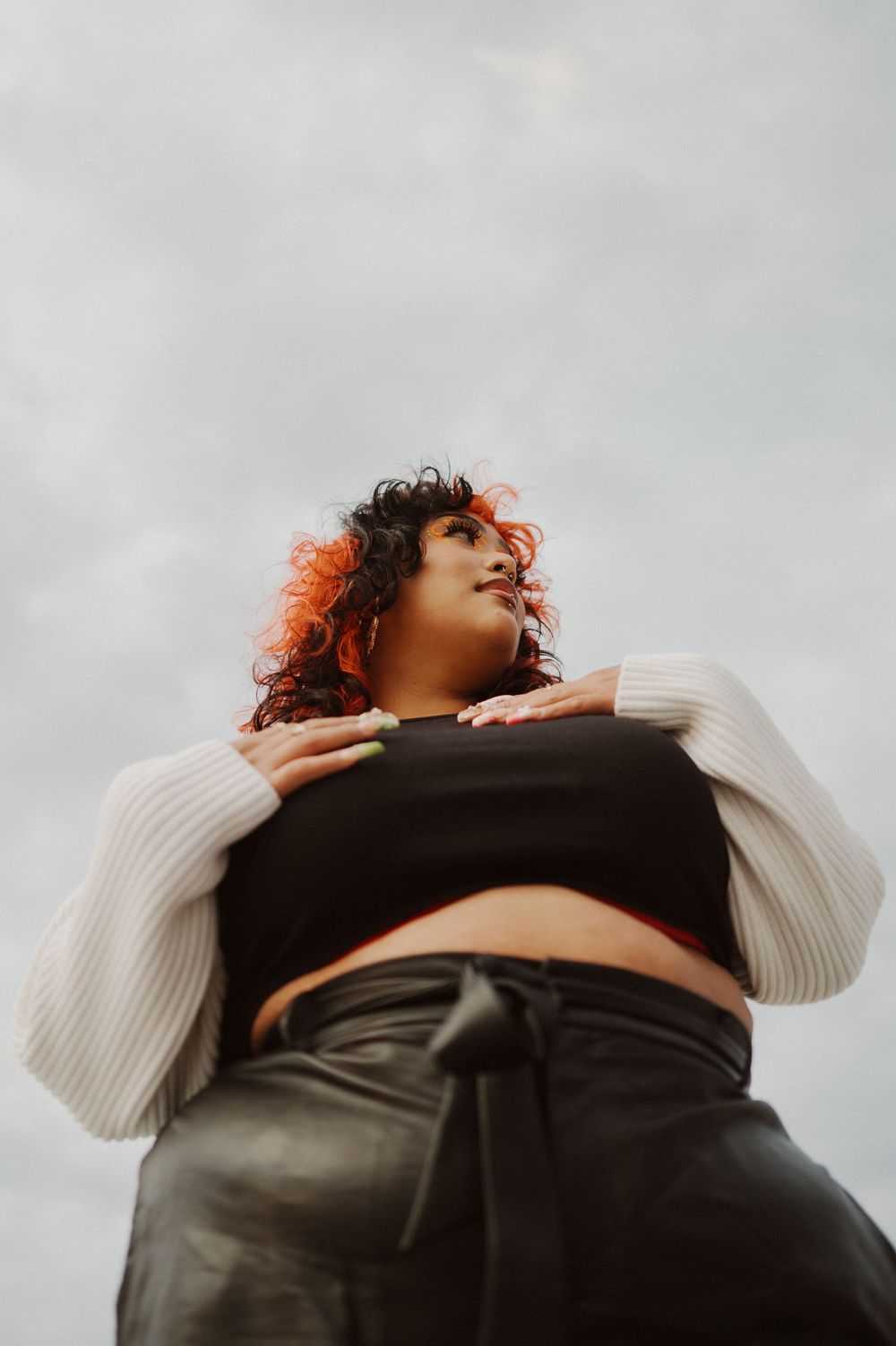 Low angle portrait against cloudy sky wearing white knit sleeves and black crop top with leather pants.
