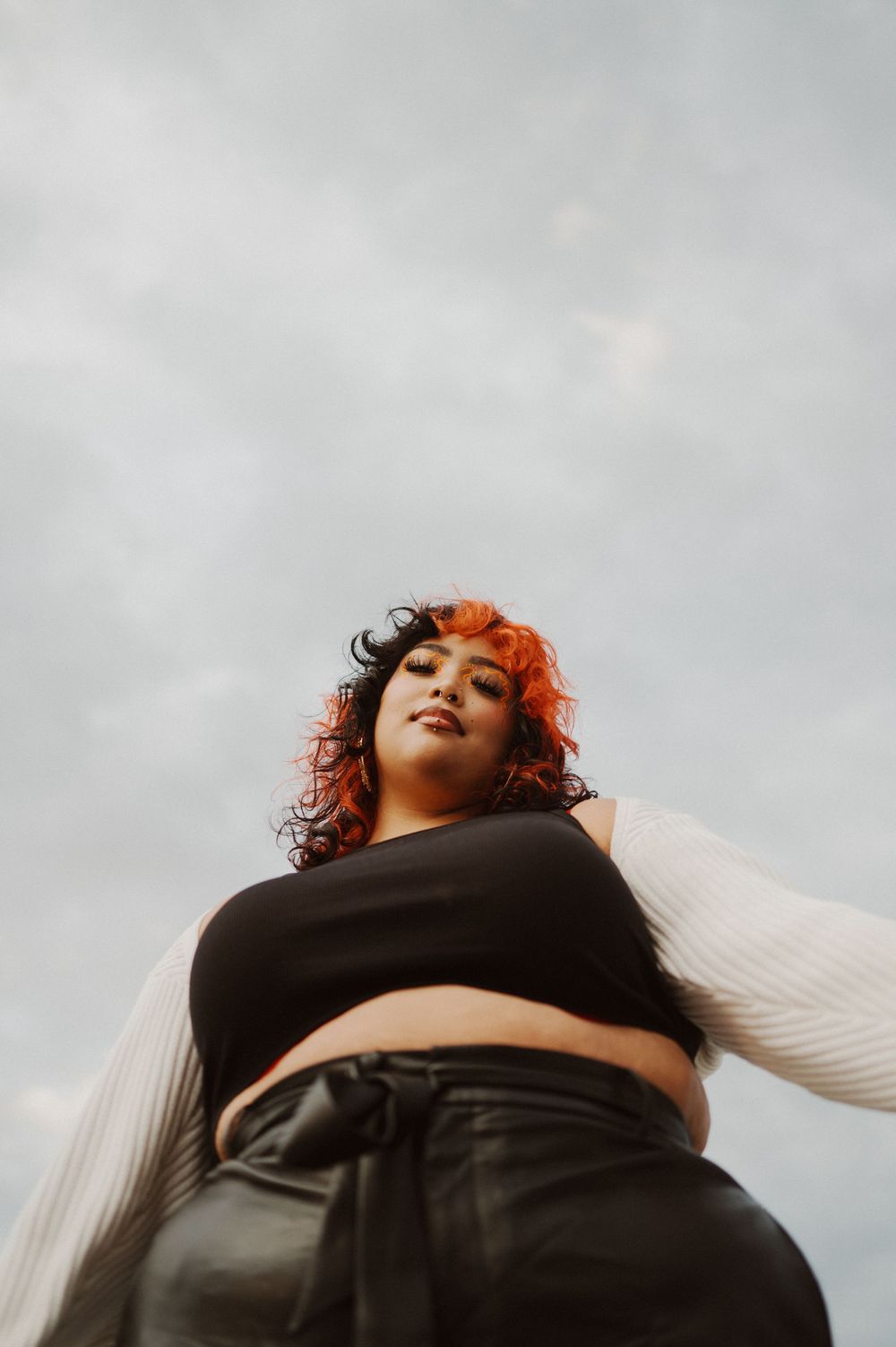 A person with curly red hair wearing a black crop top and white sleeves poses against a cloudy sky background.