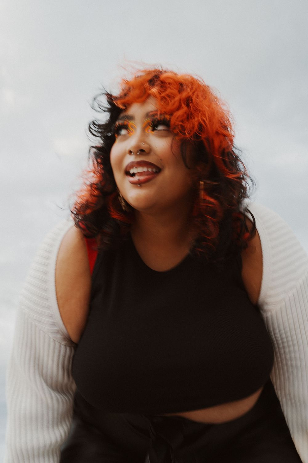 Portrait of someone with vibrant red curly hair wearing a black top and smiling against a light background.
