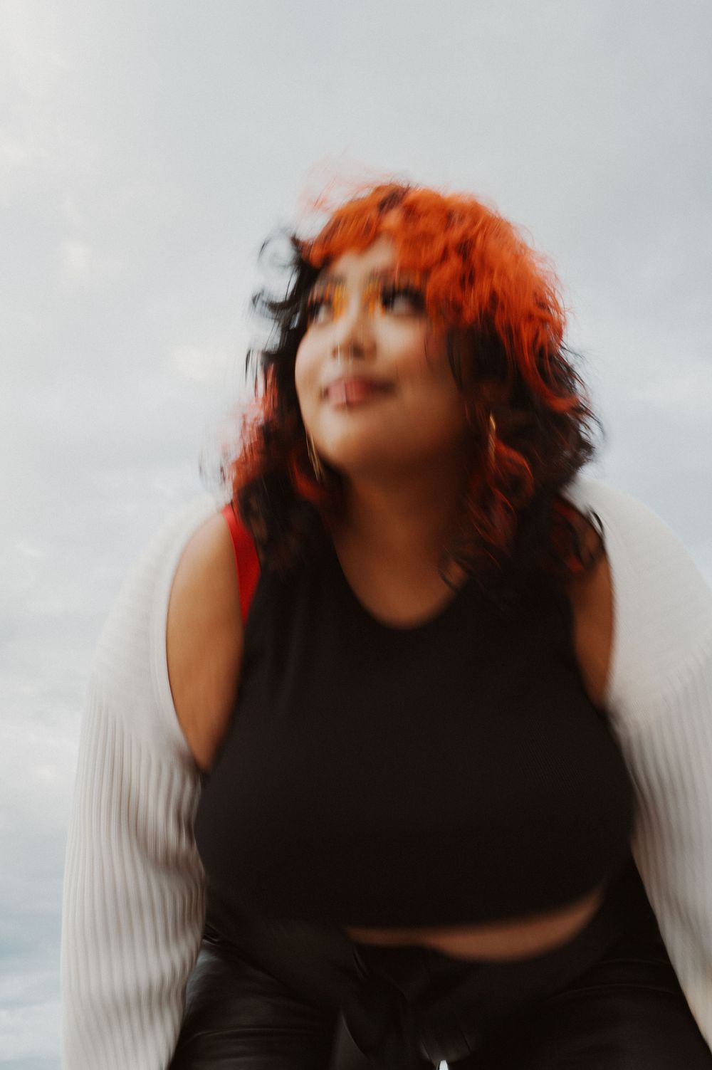 Stylized portrait with vibrant red curly hair against cloudy sky wearing black crop top in dreamy motion blur effect.